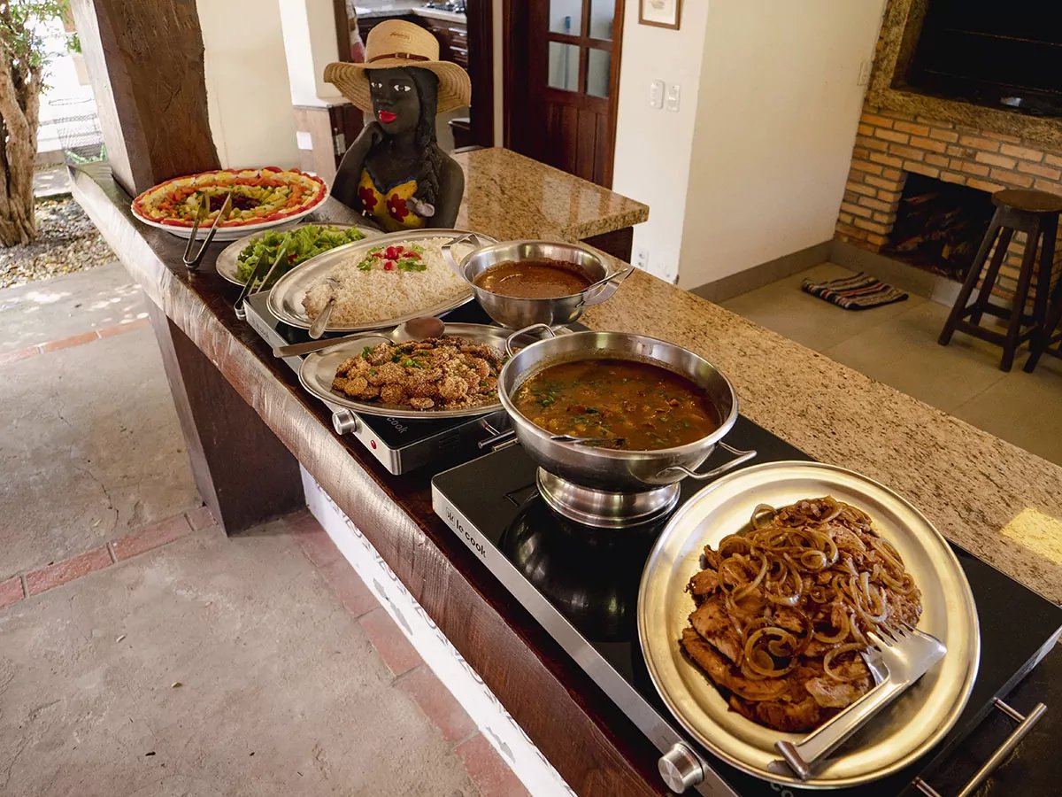 A generous lodge buffet spread on a granite counter featuring grilled meat with caramelised onions, two warm stews, rice, a fried side dish, green salad, and a colourful fruit platter — part of the full-board experience on a pantanal tour from campo grande