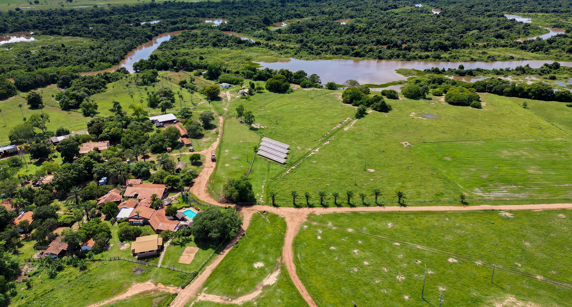 Aerial view of a Pantanal ecolodge on a pantanal tour from Campo Grande, with solar panels, red-roofed buildings, a swimming pool, dirt tracks, green pastures, and a winding river visible in the background