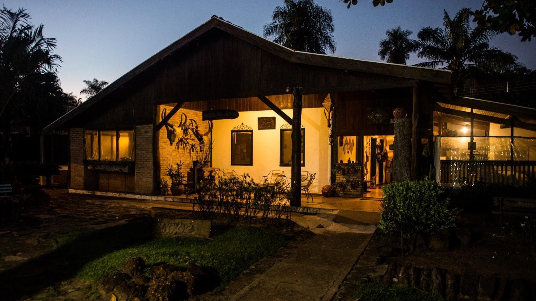 A warmly lit rustic ecolodge with a wooden gabled roof and open veranda glowing with amber light at dusk, surrounded by palm trees and tropical gardens, on a pantanal tour from campo grande