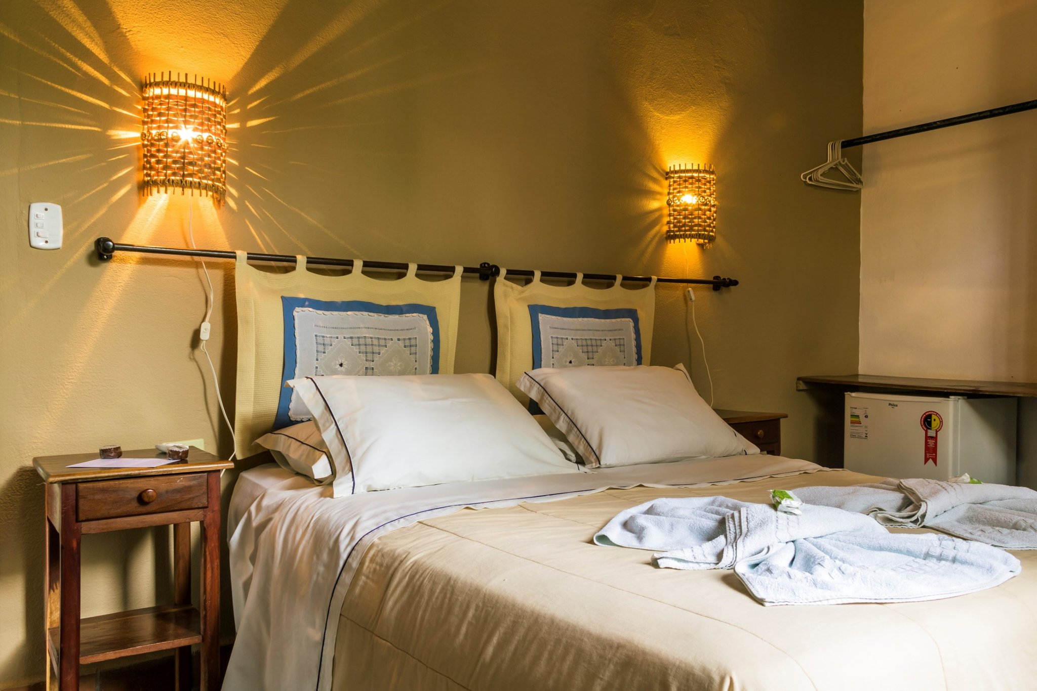 Cozy double room at a Pantanal tour from Campo Grande ecolodge, featuring warm woven wall sconces, cream bedding with decorative pillows, folded white towels and robes on the bed, a wooden nightstand, and a mini-fridge