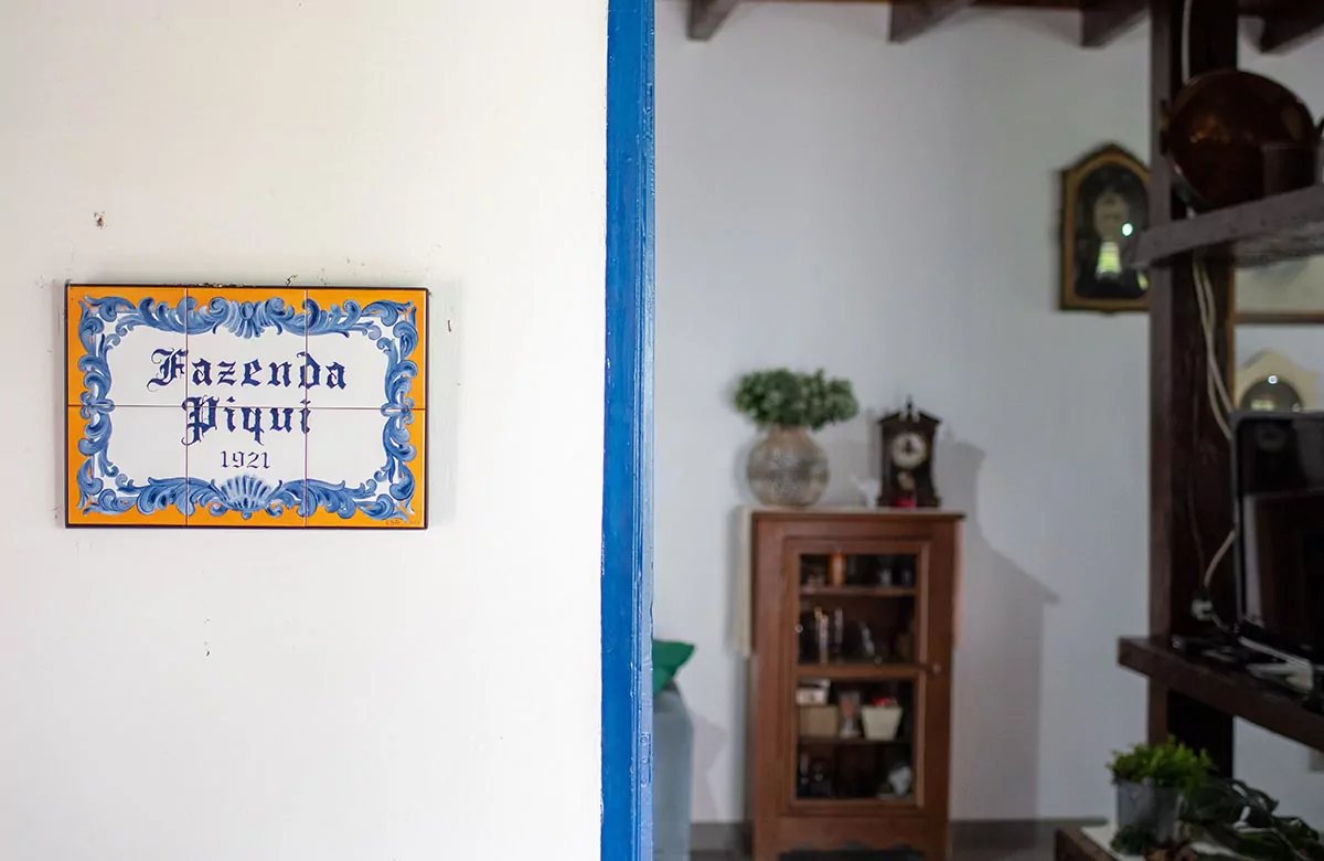 Hand-painted ceramic tile plaque reading 'Fazenda Piqui 1921' mounted on a white wall beside a blue-framed doorway leading into a rustic interior, at the start of a pantanal tour from campo grande