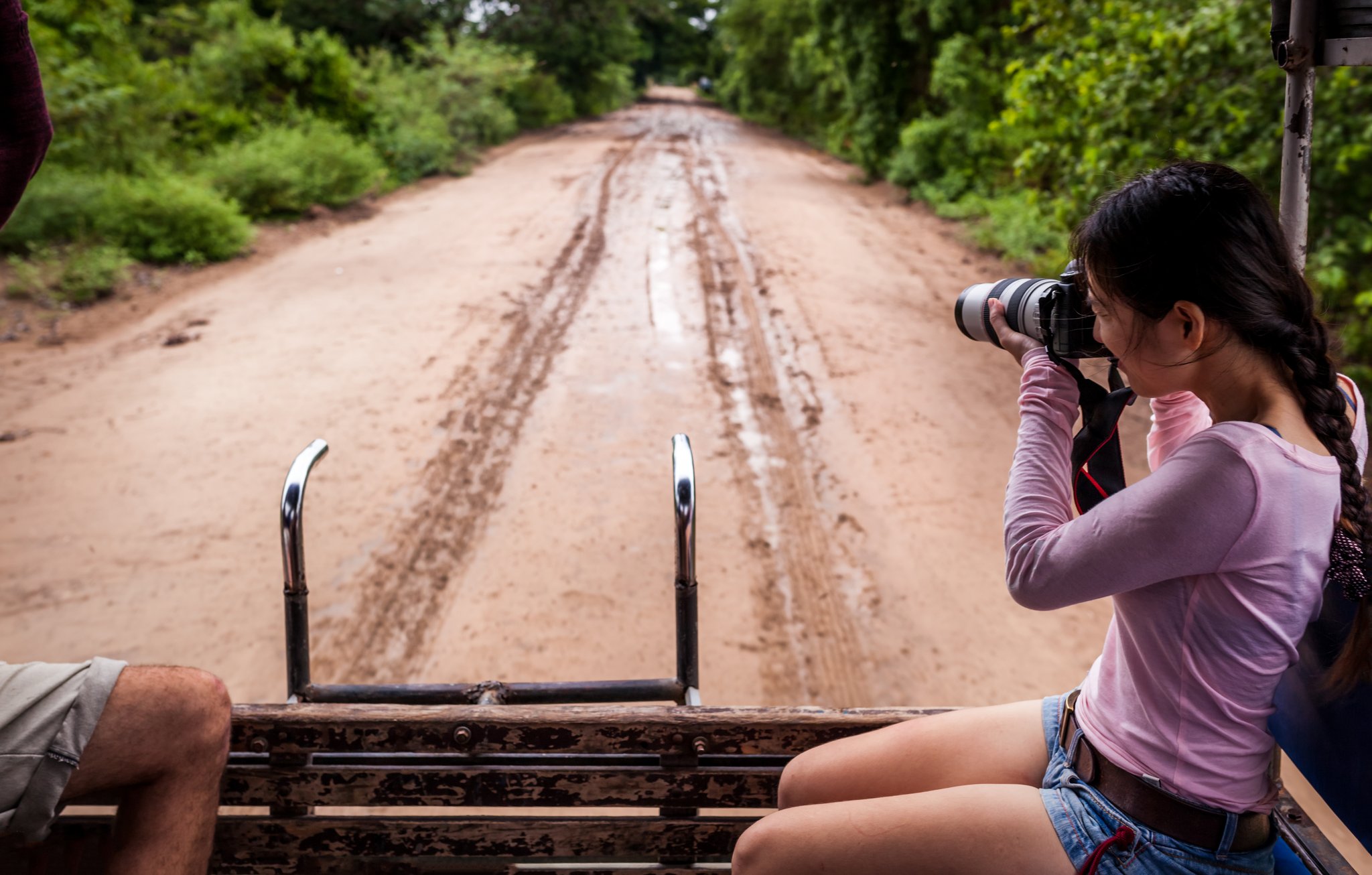 A traveller shoots with a telephoto lens from the back of an open 4x4 vehicle on a red dirt road lined with green forest during a pantanal tour from campo grande