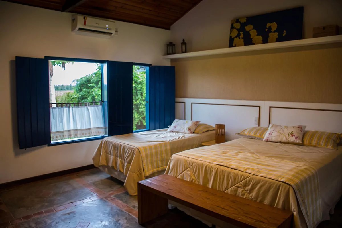 Gezellige tweepersoonskamer in een lodge met geel geruite beddengoed, terracotta tegelvloer, houten plafond en blauwe luiken die uitkijken op weelderig groen
