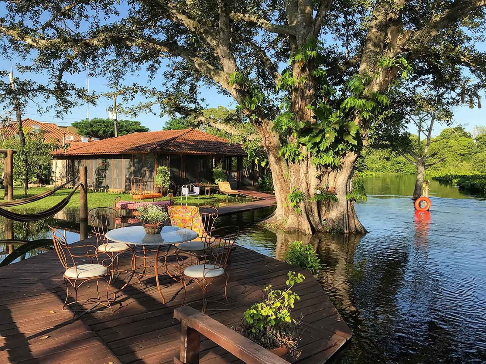 Een houten terras met smeedijzeren stoeltjes kijkt uit over kalm rivierwater; een grote boom groeit aan de oever en een ecolodge met terracotta dak is zichtbaar achter weelderig groen