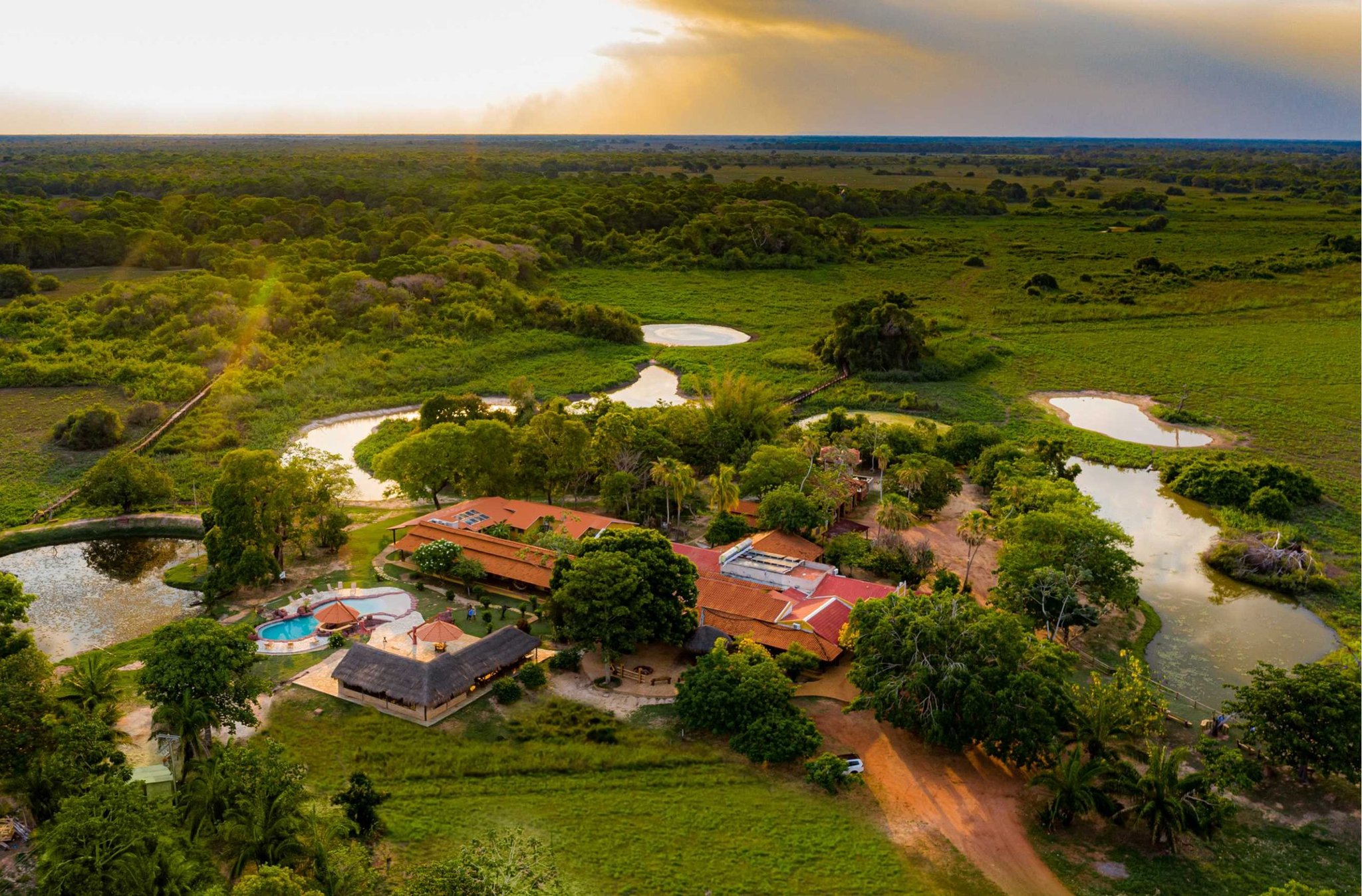 Un lodge posé au cœur des eaux, là où la faune du Pantanal vient à vous. Vue aérienne d'un éco-lodge du Pantanal entouré de lagunes sinueuses, d'une végétation de zones humides et d'un bâtiment principal aux toits en terre cuite avec piscine, au coucher de soleil doré