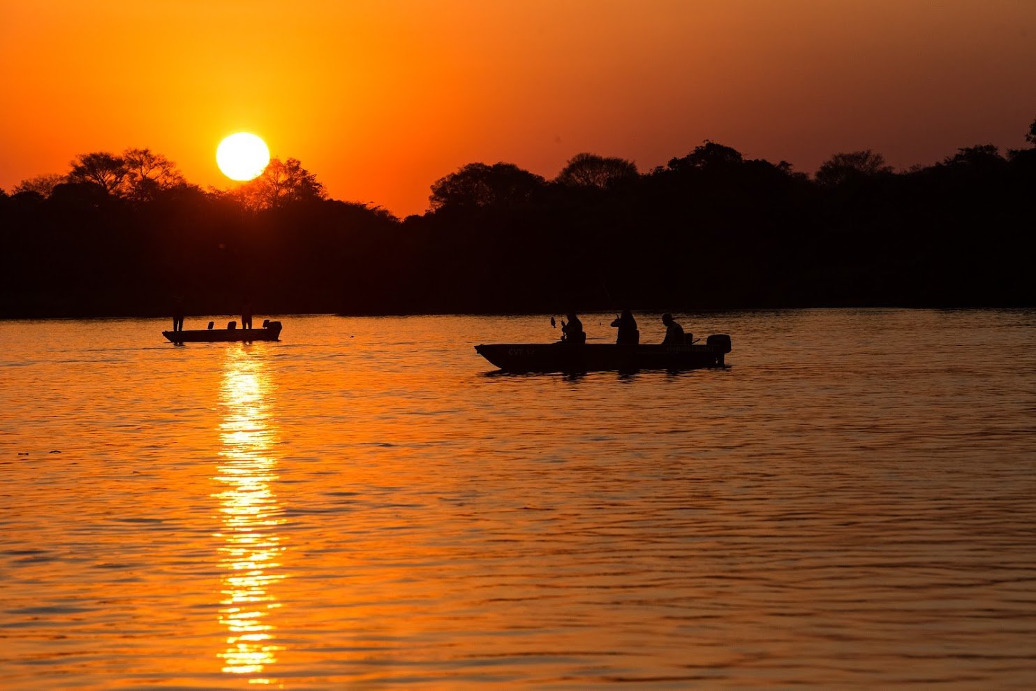 Two small fishing boats silhouetted against a blazing orange sunset over a calm river on a Pantanal Tour from Cuiabá, with a dark treeline reflected in the glowing water