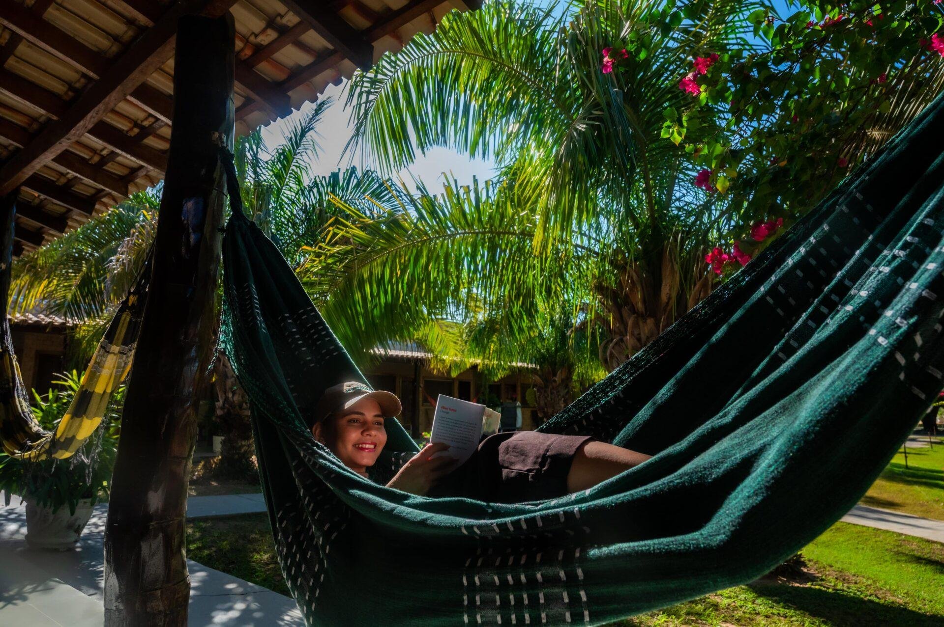 A smiling traveller reads a book while resting in a dark green hammock on a shaded lodge veranda, surrounded by palm fronds and pink bougainvillea, during a Pantanal Tour from Cuiabá