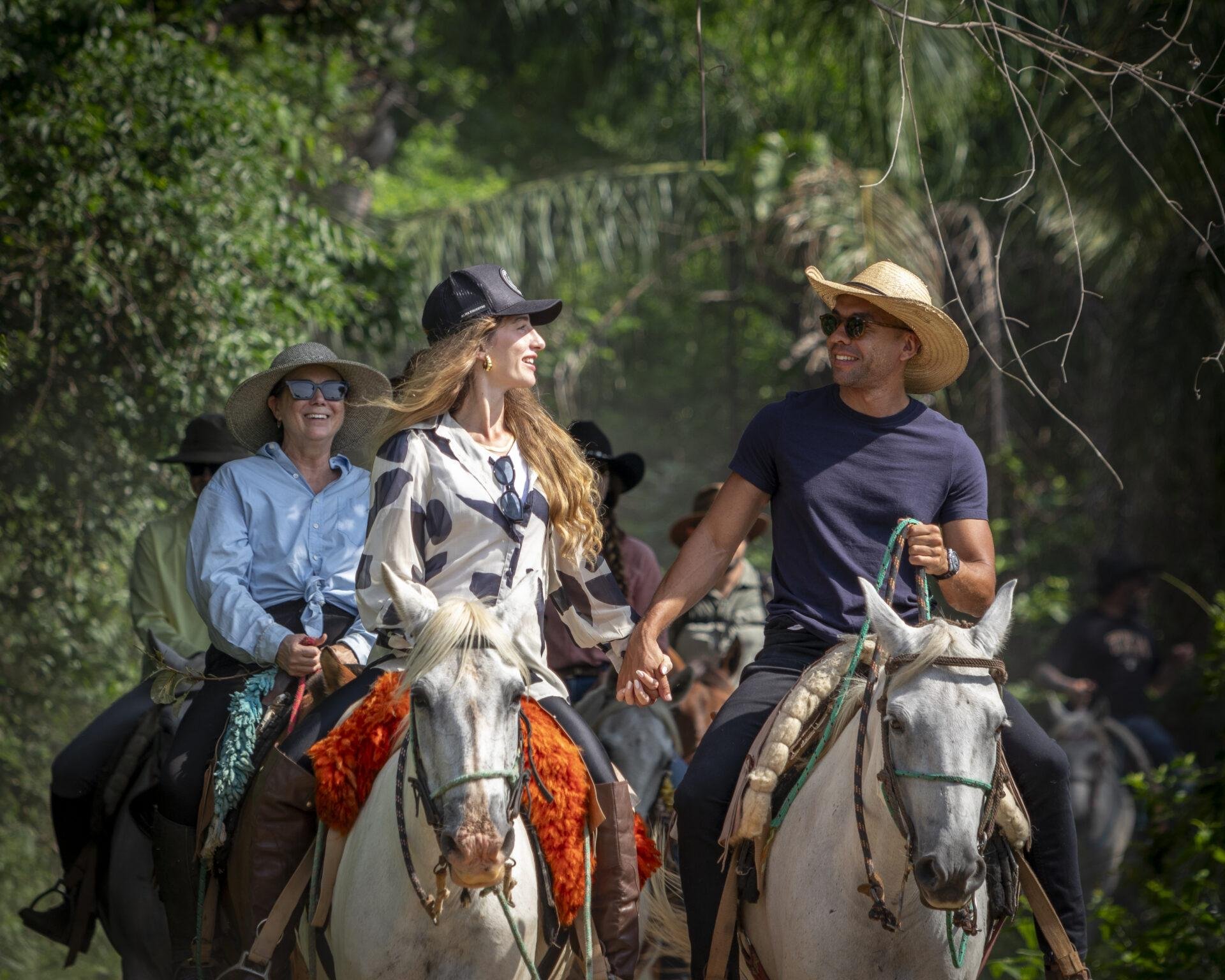 A group of smiling riders on grey horses moves through a lush green corridor on a Pantanal Tour from Cuiabá, a couple in the foreground holding hands in the saddle