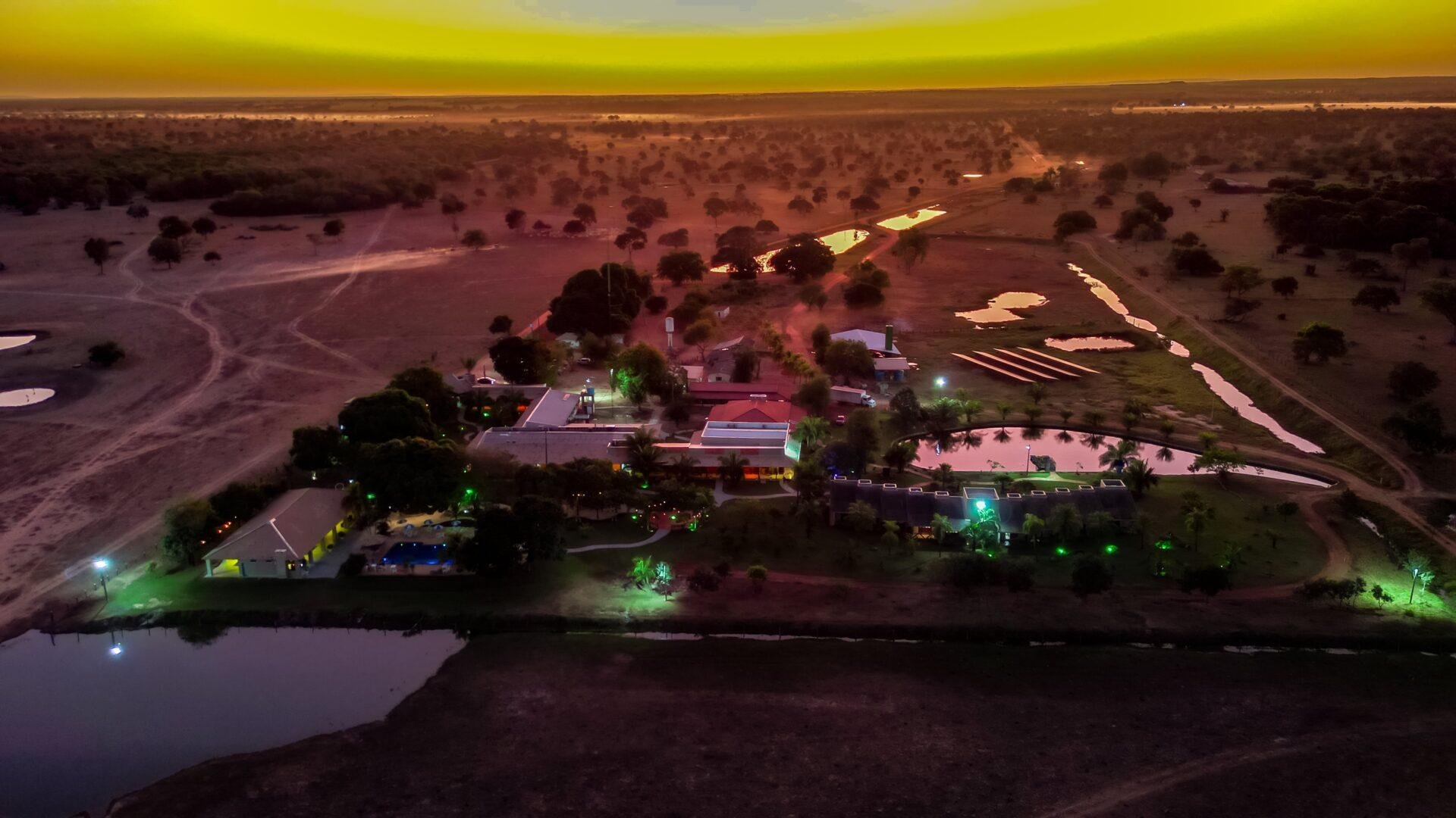 Aerial drone view of an illuminated Pantanal lodge at dusk, surrounded by wetland ponds and open savanna scrubland stretching to the horizon under a vivid yellow-orange sky — a base for a Pantanal Tour from Cuiabá