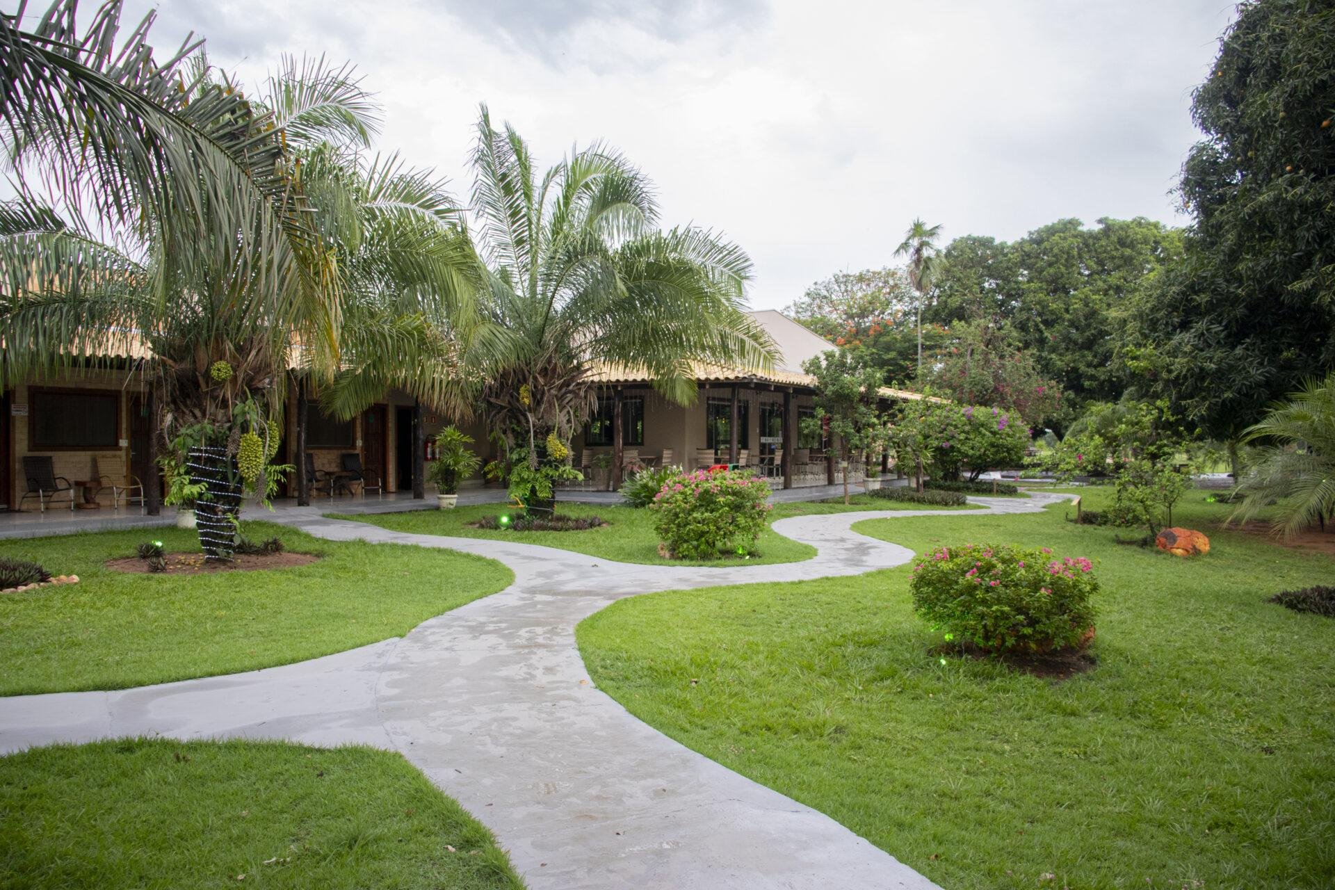 Winding concrete pathways curve through a lush tropical garden leading to low-slung thatched-roof lodge buildings on a Pantanal Tour from Cuiabá, with palm trees, flowering shrubs, and manicured lawns under a cloudy sky