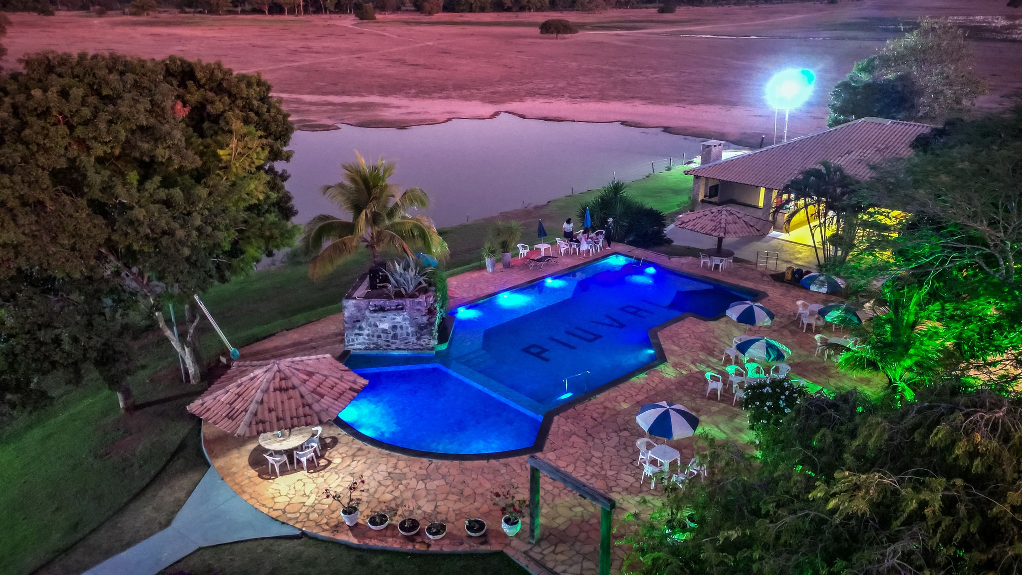 Aerial night view of an illuminated swimming pool at a Pantanal Tour from Cuiabá lodge, with patio umbrellas, a stone water feature, and a natural lake stretching across open savanna in the background
