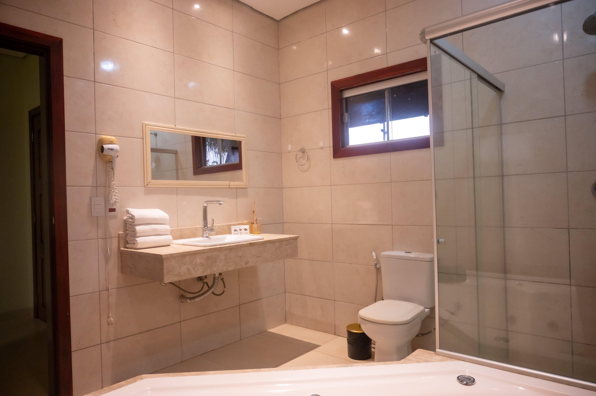 Spacious private bathroom at a Pantanal Tour from Cuiabá lodge, featuring a marble-finish vanity, folded white towels, glass-enclosed shower, bathtub, and a wall-mounted hair dryer