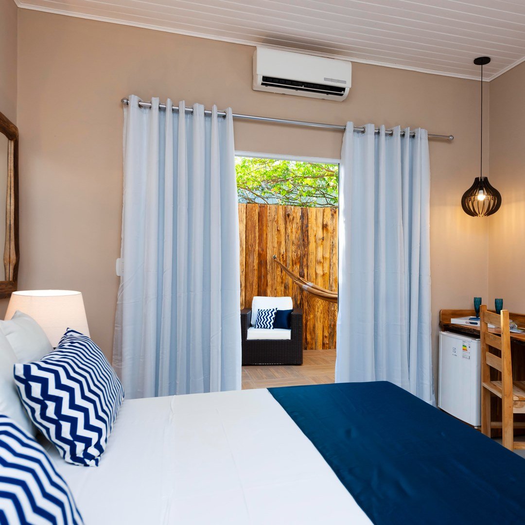 Air-conditioned double room on a Pantanal Tour from Cuiabá, with navy chevron pillows, white curtains, and a private wooden terrace with hammock and lounge chair visible through the open sliding door