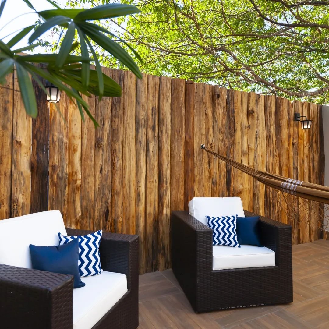 Shaded terrace at a Pantanal Tour from Cuiabá lodge, with dark rattan armchairs, white cushions, blue chevron pillows, a hammock, and rustic timber walls beneath green tree canopy