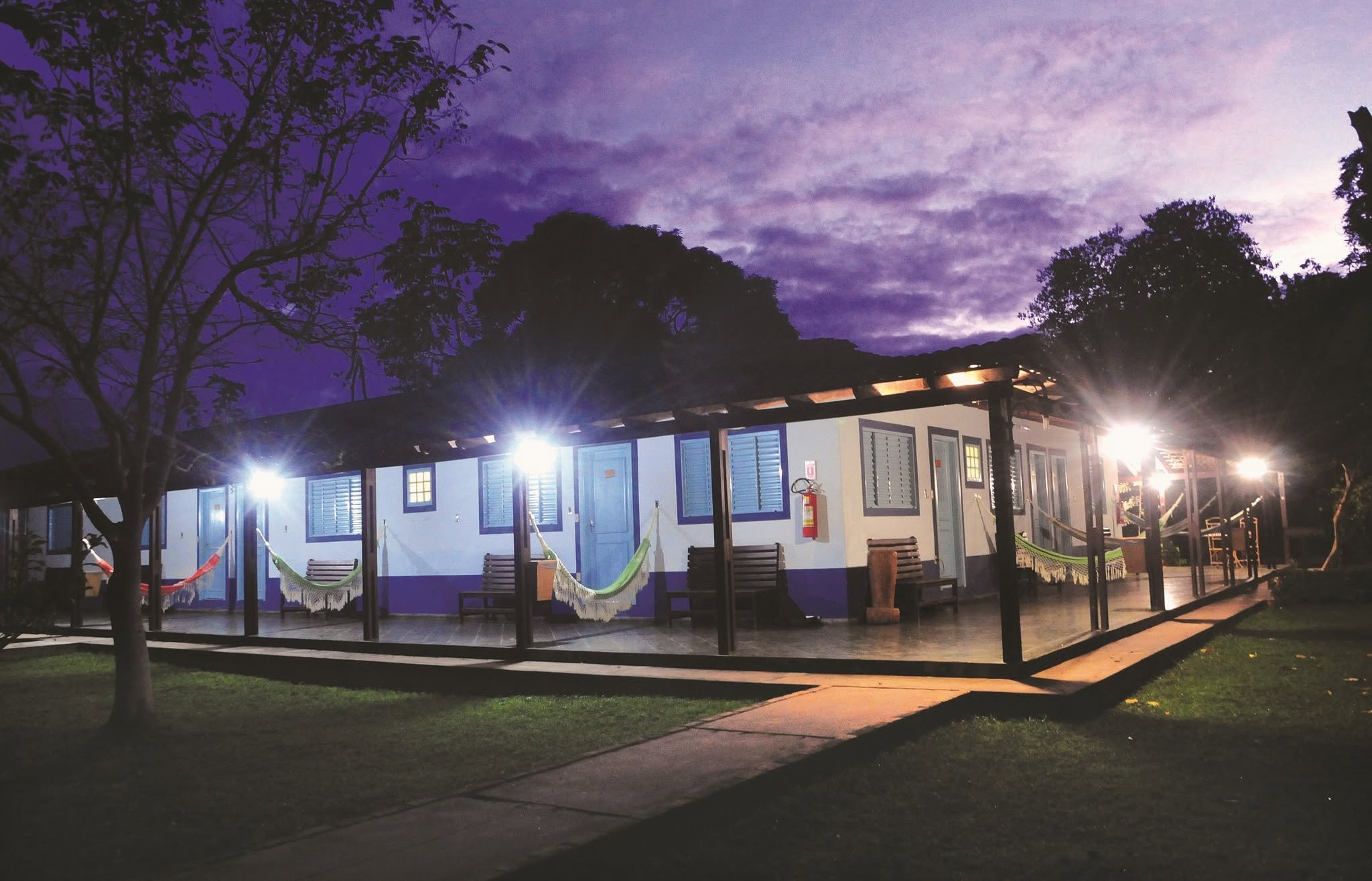 A low white lodge building with colorful hammocks strung along a covered veranda, glowing under bright spotlights against a deep purple twilight sky on a Pantanal Tour from Cuiabá