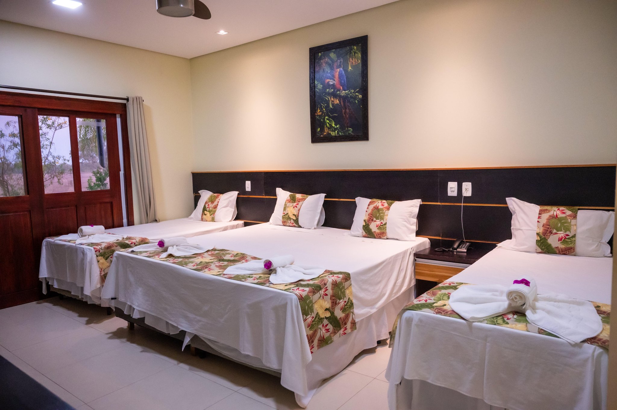 A triple-occupancy lodge room on a Pantanal Tour from Cuiabá, with three neatly made beds featuring tropical floral runners, rolled towels with purple orchid accents, and a framed macaw painting above the headboard