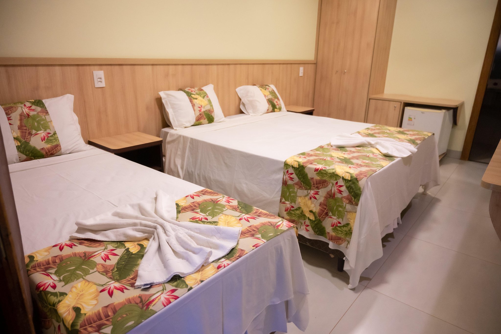 Three neatly made beds with tropical floral bedspreads and folded white towels in a clean, wood-panelled guest room on a Pantanal Tour from Cuiabá