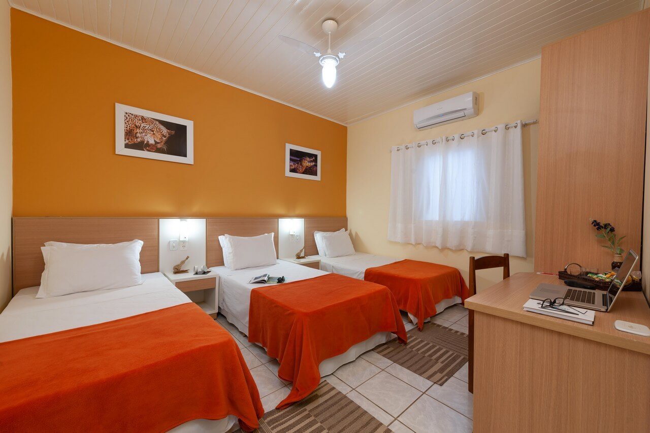 A bright triple room at a Pantanal Tour from Cuiabá lodge, featuring three single beds with orange throws, a warm amber accent wall adorned with wildlife prints, air conditioning, and a work desk with a laptop
