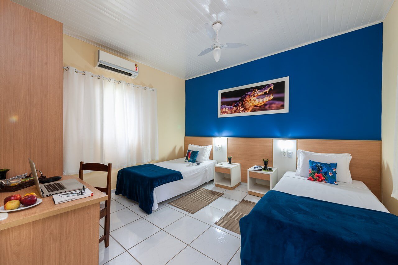 Twin room at a Pantanal Tour from Cuiabá lodge, featuring two beds with navy throws, a bold blue accent wall, a framed caiman photograph, and a work desk with fresh fruit