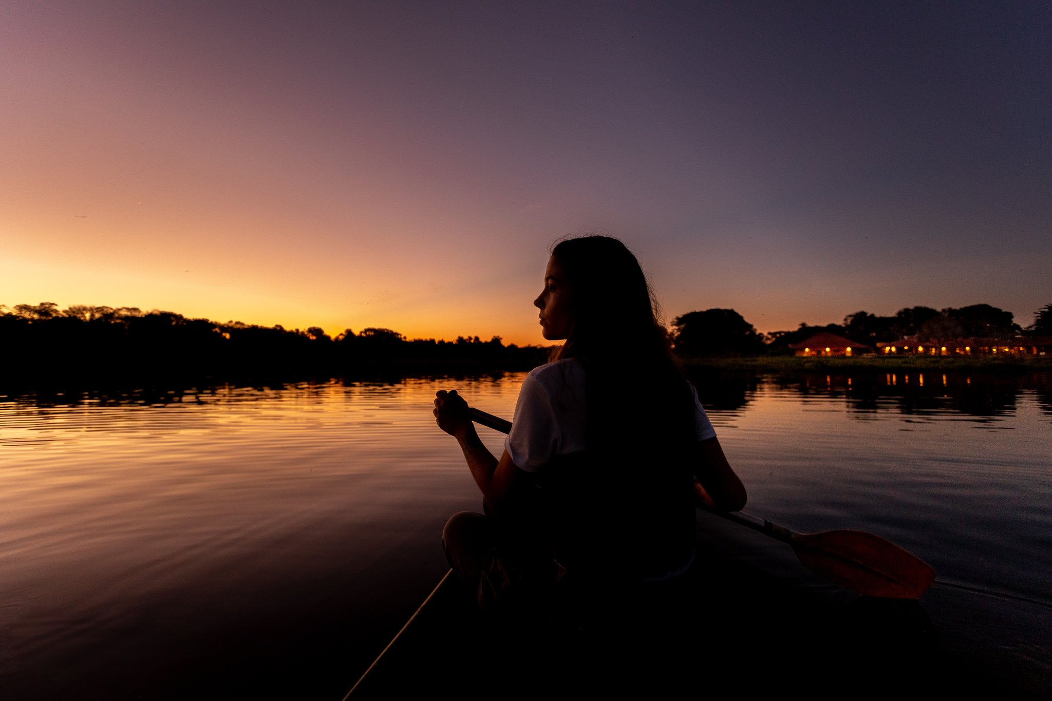 Silhueta de uma mulher em caiaque em um rio tranquilo do Pantanal Sul ao entardecer, com o céu em tons de laranja e roxo e as luzes de uma lodge ao fundo