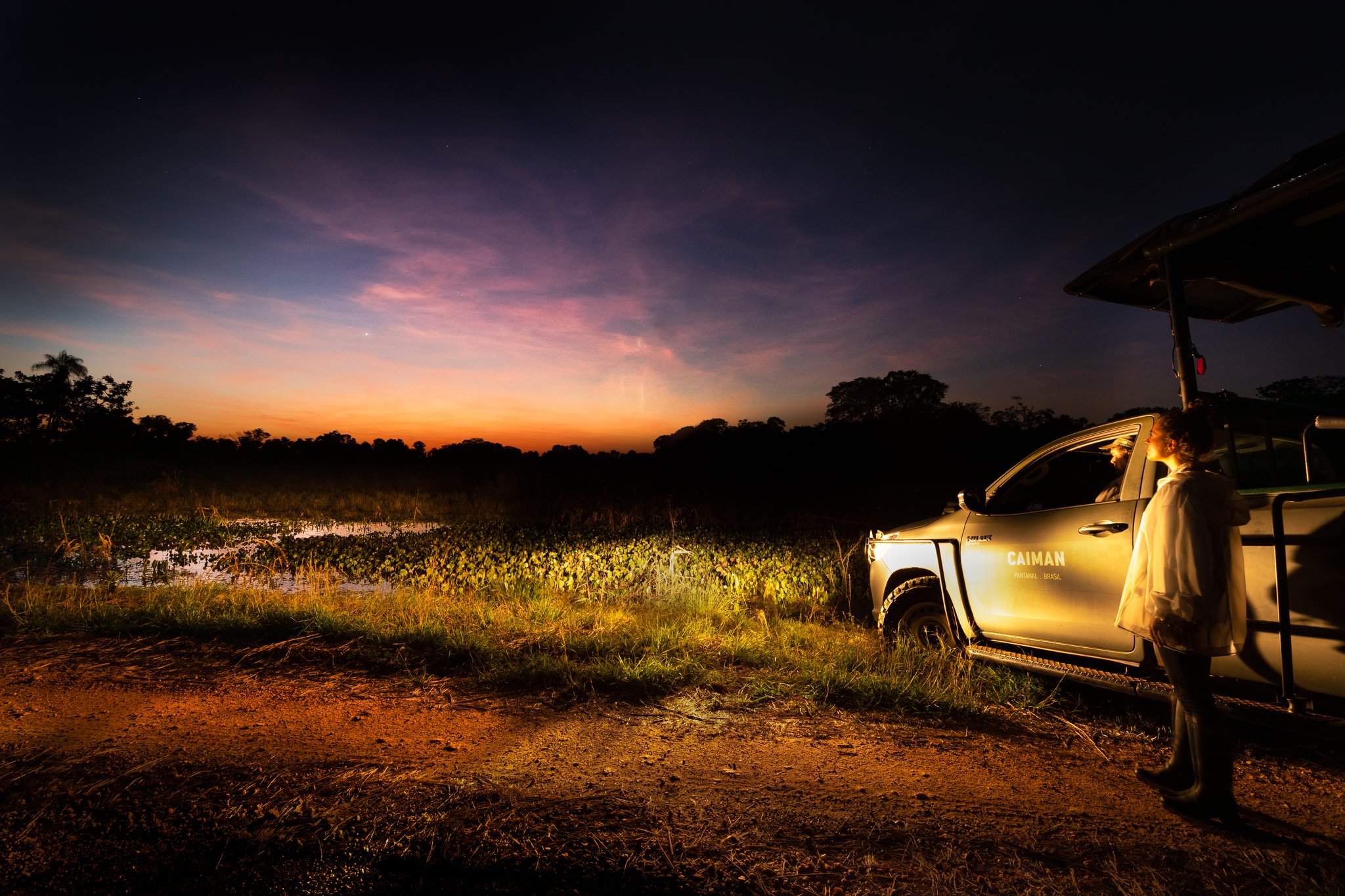 Um veículo da Caiman Refugio Ecológico estacionado às margens de um brejo alagado ao entardecer durante um passeio noturno pelo Pantanal no Brasil, com um guia iluminado por luz artificial sob um céu alaranjado e roxo