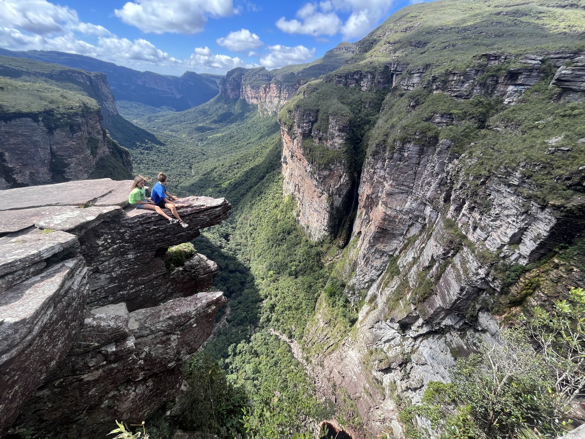Two tiny figures, one vast canyon — the Cachoeirão lookout earns every step. Two hikers sitting on a sheer rock ledge on the Pati Valley hike, gazing over a vast canyon with vertical sandstone cliffs and a forested valley stretching to the horizon under a blue sky