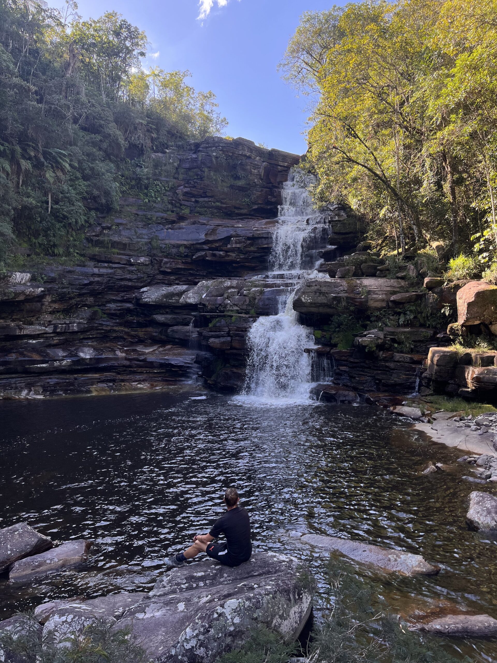 Sit, breathe, let the falls do the talking. A hiker sits alone on a flat sandstone rock beside a dark natural pool, gazing up at a tiered waterfall cascading down layered rock faces during the Pati Valley hike, surrounded by lush green forest under a clear blue sky