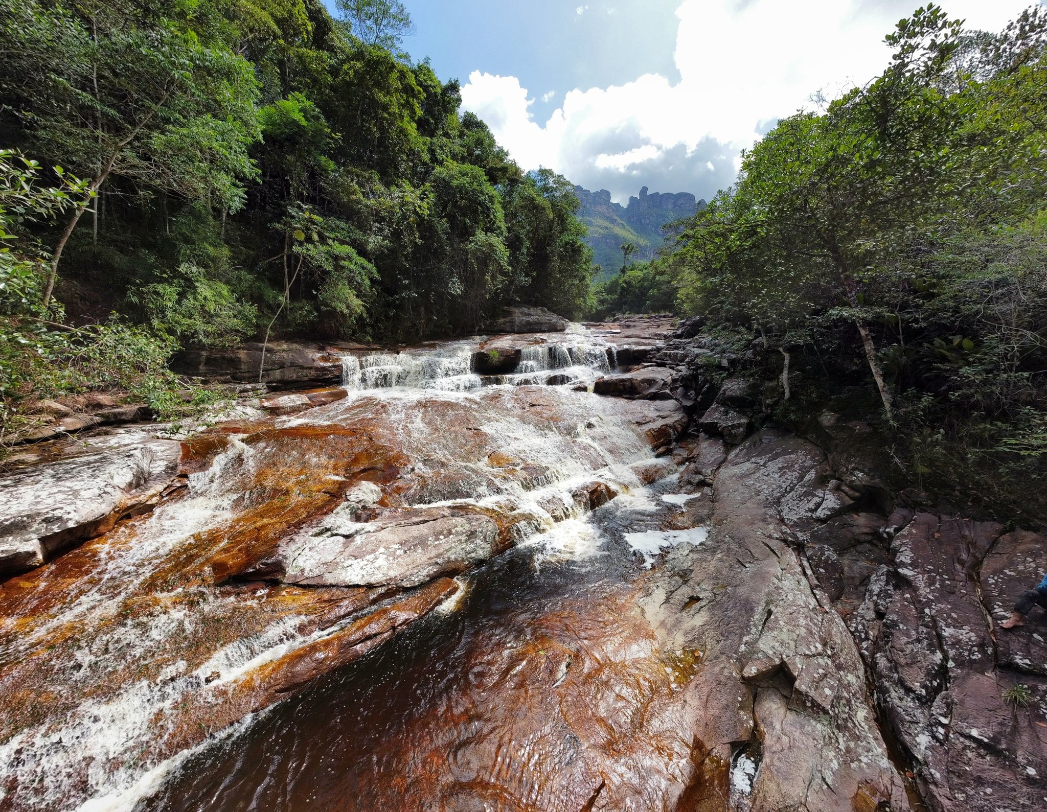Layer after layer, the water finds its way down. Clear water rushing over rust-stained sandstone ledges on the Pati Valley hike, with dense forest and distant rocky peaks rising under a cloudy sky