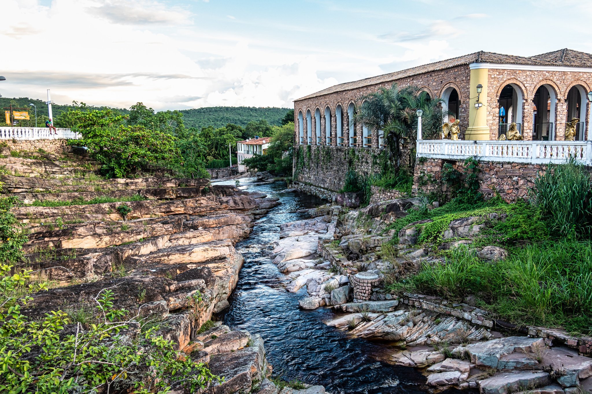 A stone colonial building with arched windows overlooks a clear river cutting through flat sandstone rocks in a Brazilian highland town, the gateway to the Pati Valley hike