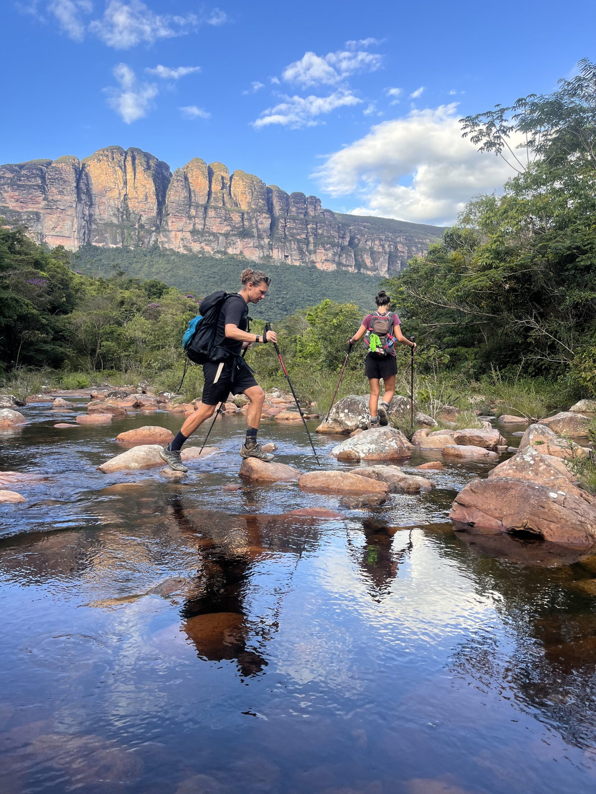 Stepping stone by stone, the canyon walls watching over every move. Photo: Lucas Neves Two hikers with trekking poles stepping across rounded rocks in a shallow river on the Pati Valley hike, with dramatic sandstone cliffs rising behind the green forest under a blue sky