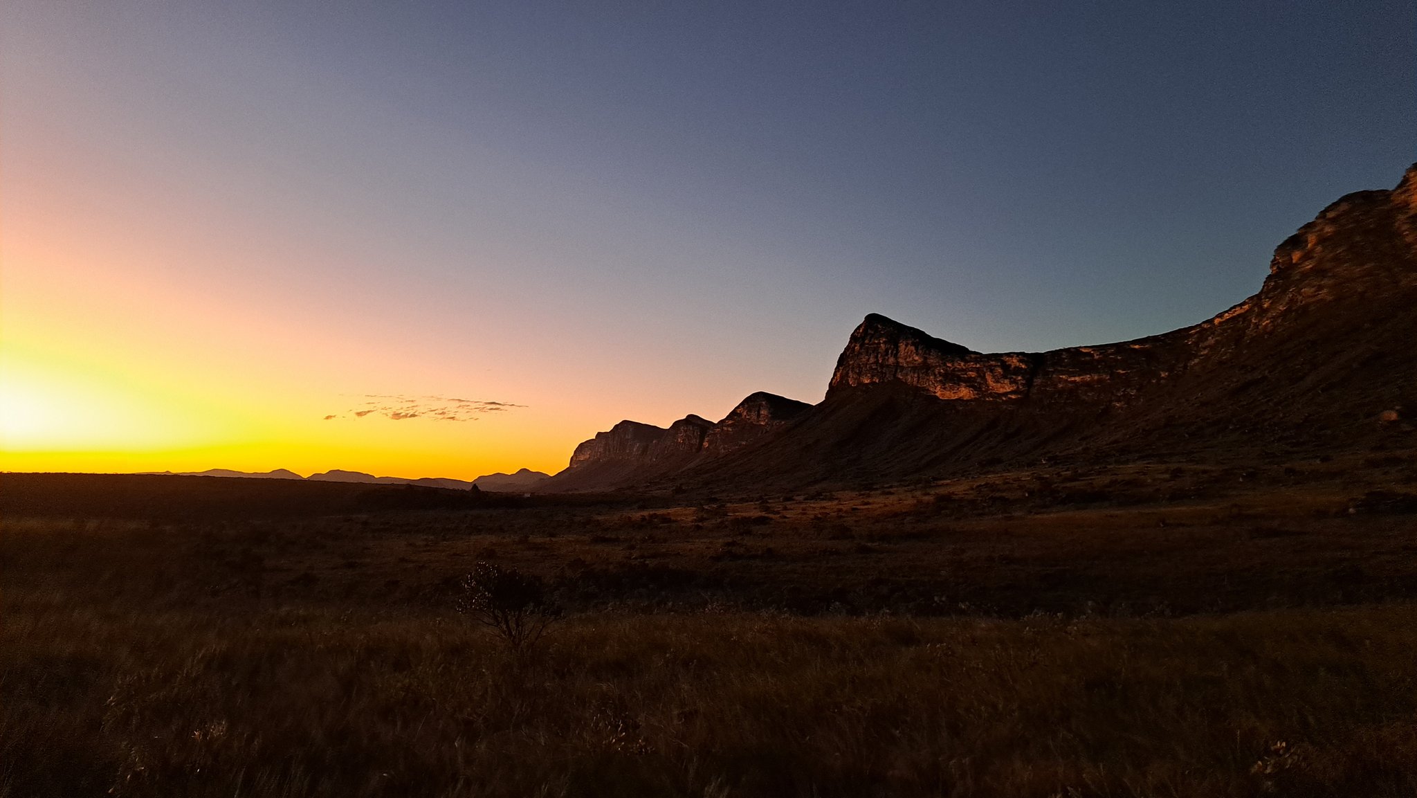 Last light catching the cliffs — the valley settles into silence. A vivid orange and yellow sunset glows on the horizon behind the silhouette of a rocky escarpment during the Pati Valley hike, with open savanna grassland in the foreground