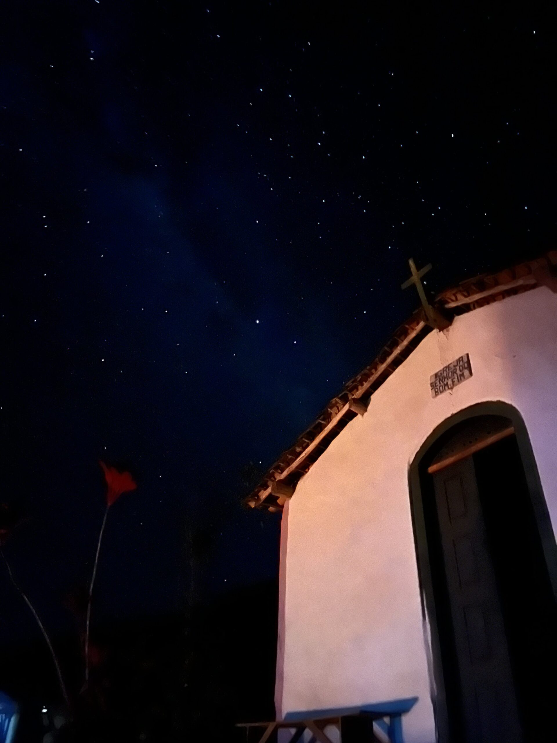 Stars thick overhead, a rooftop cross holding its own against the night. A whitewashed colonial chapel signed 'Igreja Senhor do Bonfim' glows under a star-filled night sky on the Pati Valley hike, with a rooftop cross silhouetted against the Milky Way
