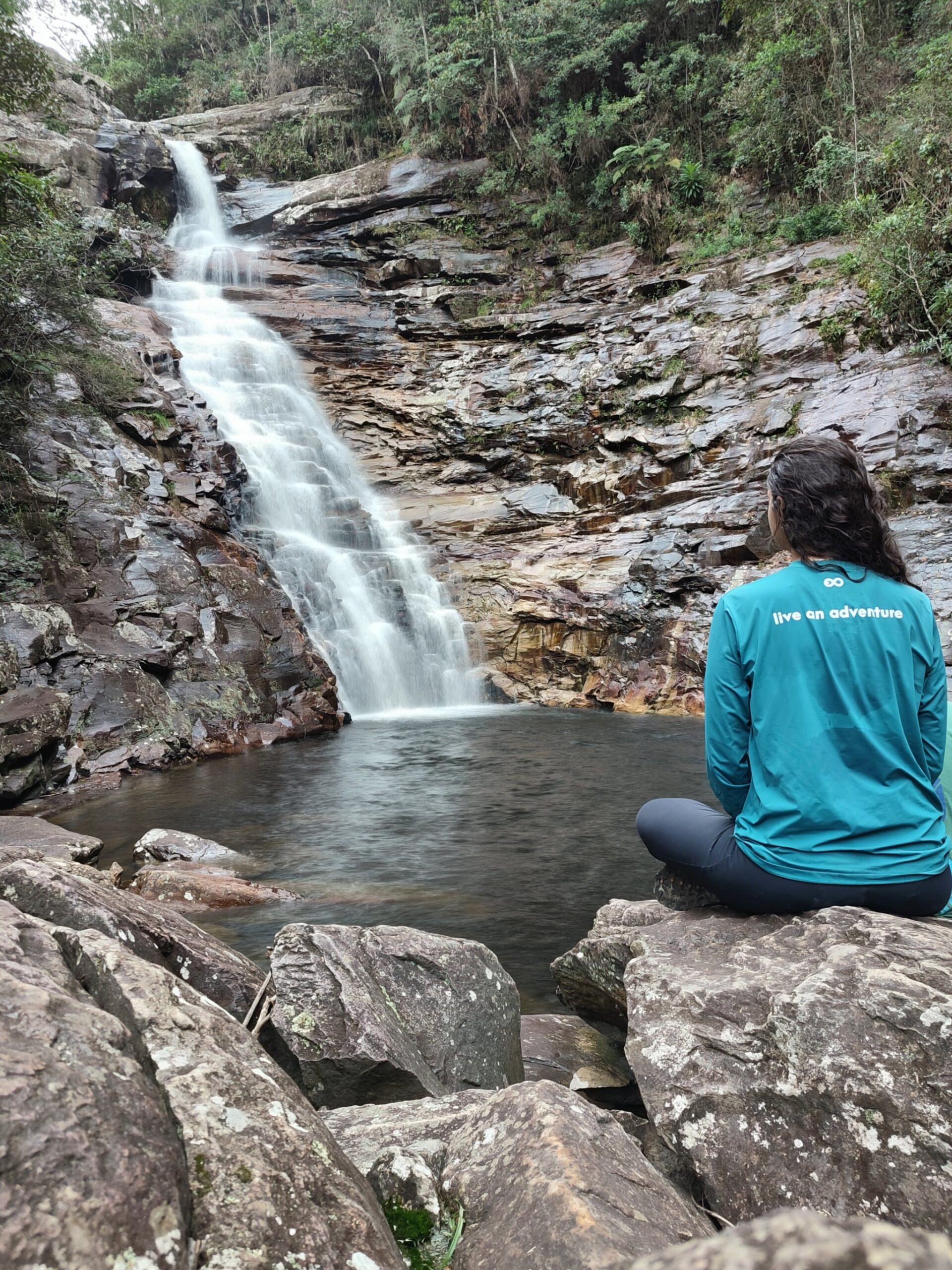 Still mind, moving water — a quiet moment on the trail. A traveller in a teal top sits cross-legged on rocks during the Pati Valley hike, gazing at a silky waterfall cascading into a still pool surrounded by layered stone cliffs and forest