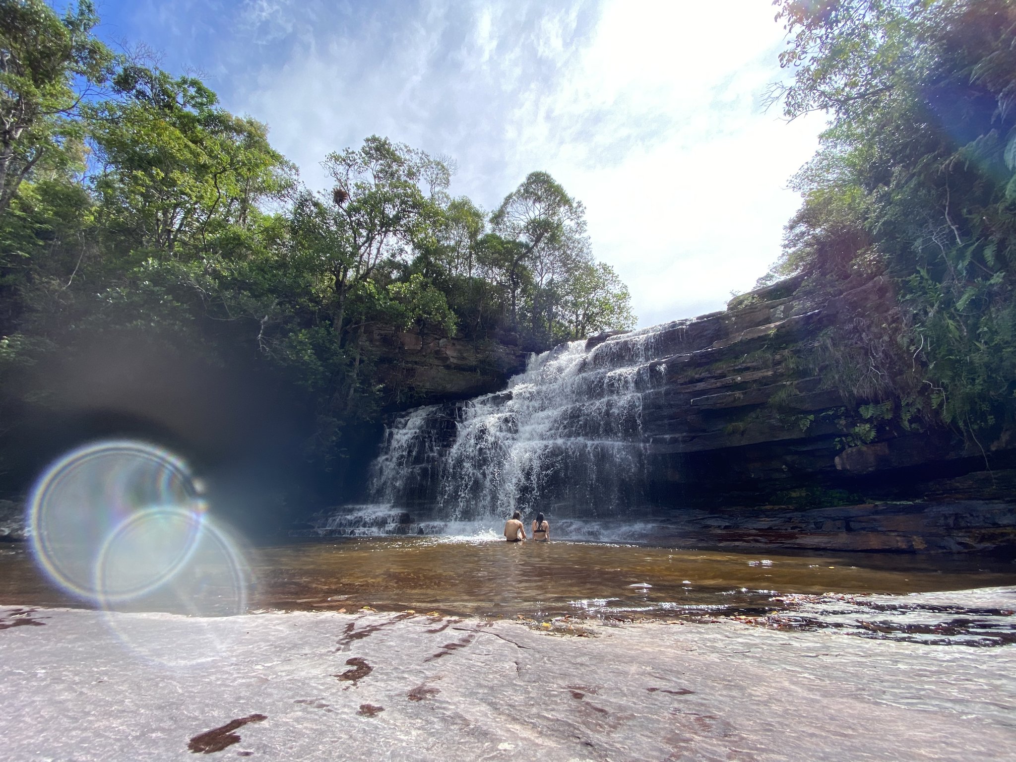 Two swimmers wading in a wide natural pool beneath a multi-tiered rock waterfall on the Pati Valley hike, surrounded by green cerrado trees under a bright sky
