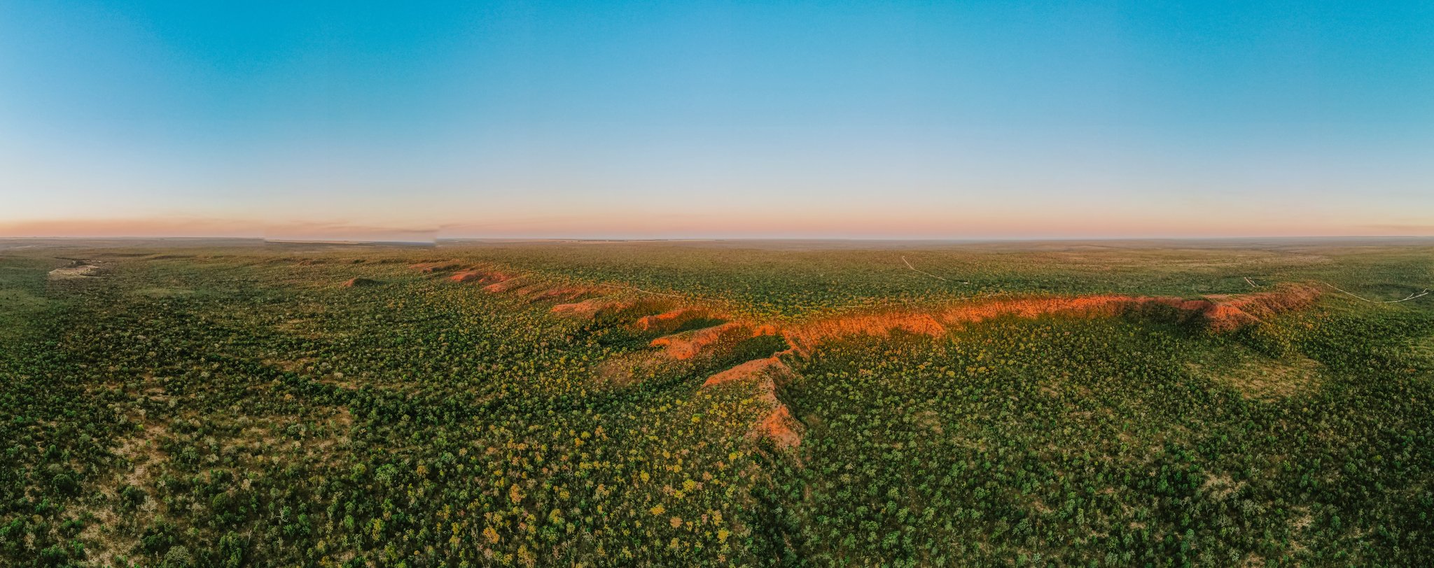 The Cerrado runs to the horizon, and that iron-red ridge reminds you the land has its own ideas about colour. Aerial panoramic view of Pousada Trijunção's vast Cerrado landscape at sunset, with a vivid red laterite ridge cutting through dense green savanna under a pastel pink and blue sky