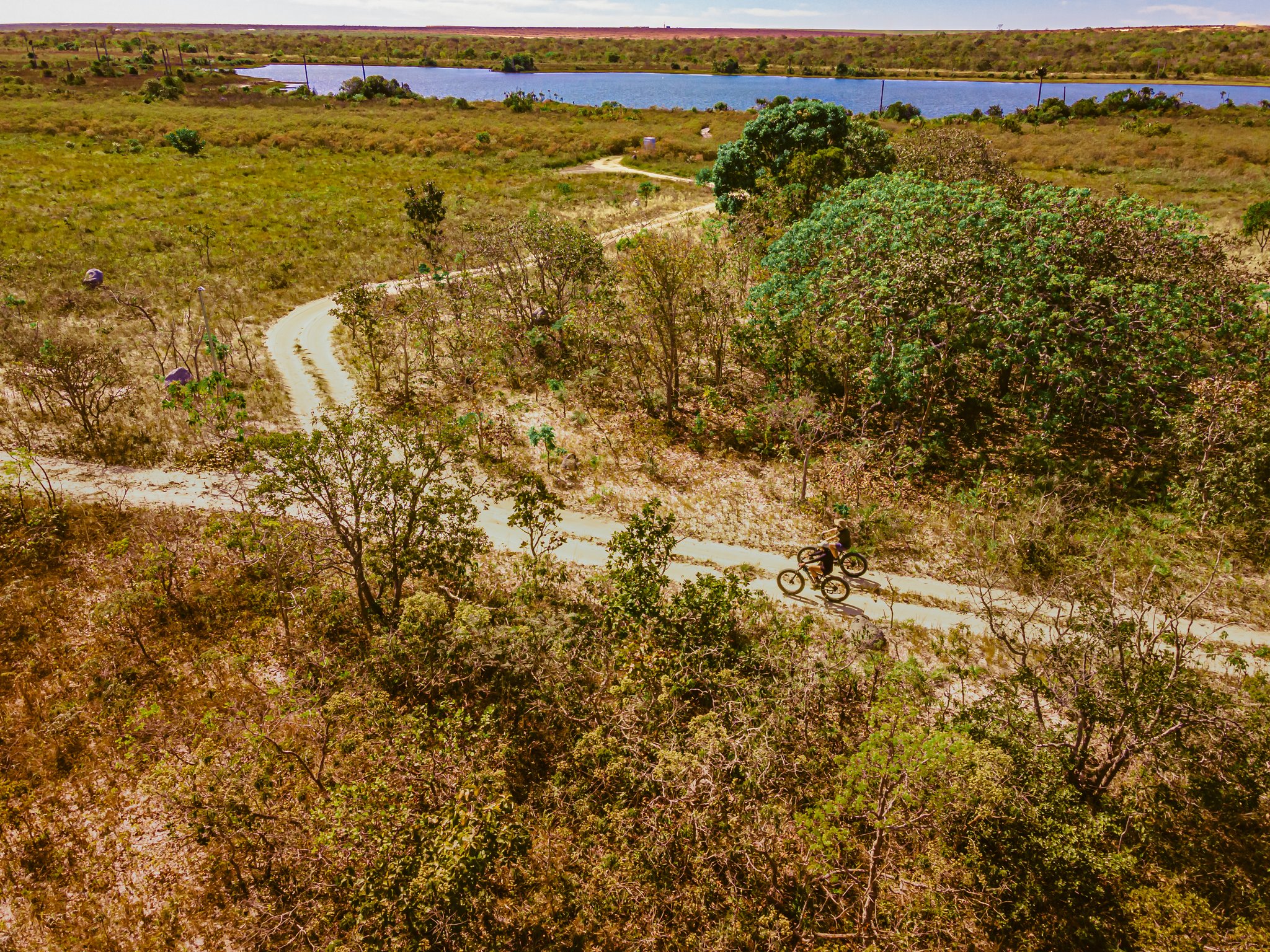 Two wheels, endless Cerrado — fat biking the trails of Trijunção. Aerial view of two cyclists riding fat bikes along a winding dirt trail through Cerrado scrubland at Pousada Trijunção, with a blue lagoon visible in the background