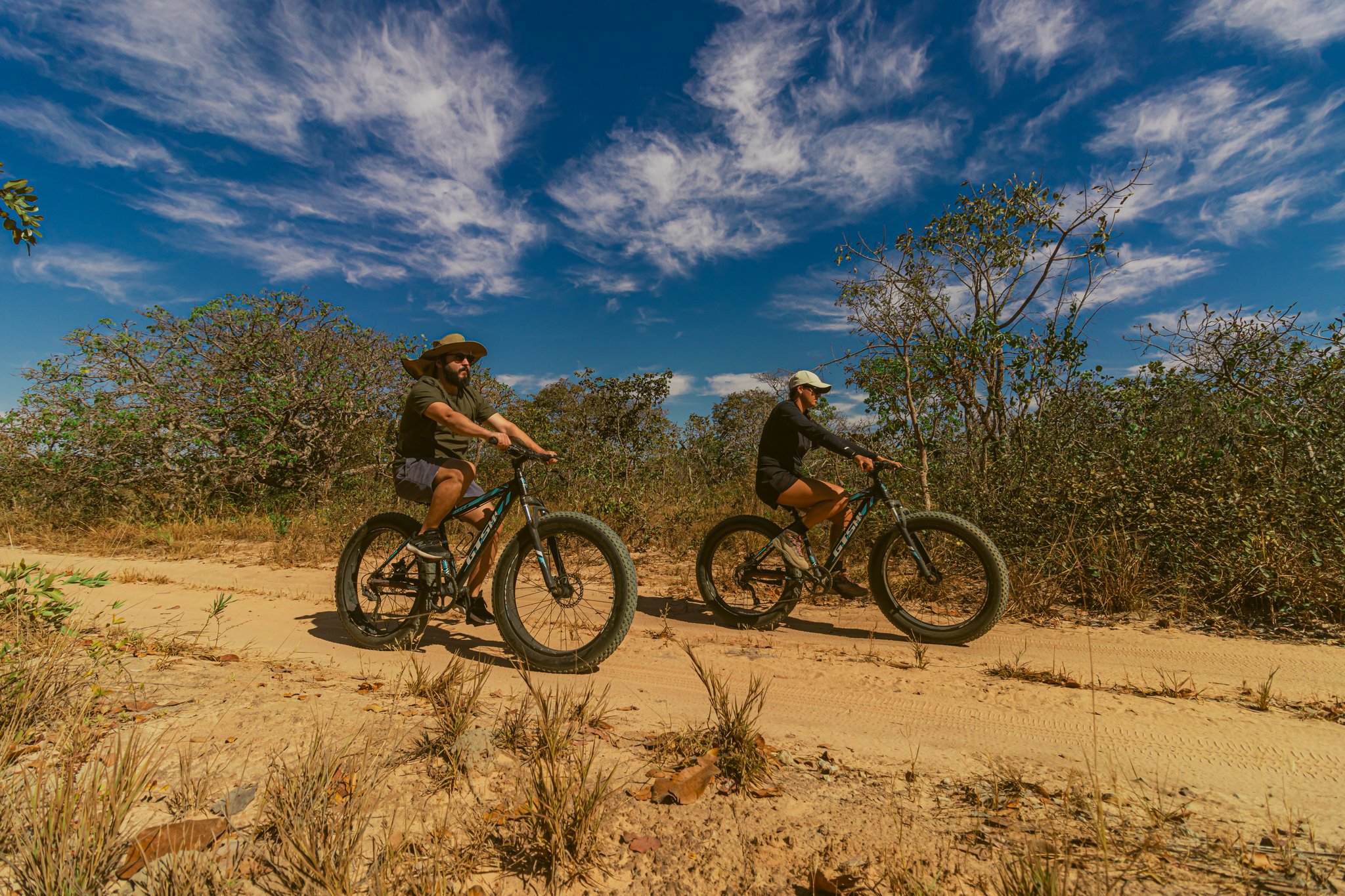Pedalling through the wild heart of the Cerrado on fat-tyre bikes built for sand and savanna. Photo: Albori Ribeiro Two cyclists riding fat bikes along a sandy Cerrado trail at Pousada Trijunção, surrounded by native cerrado shrubs under a vivid blue sky