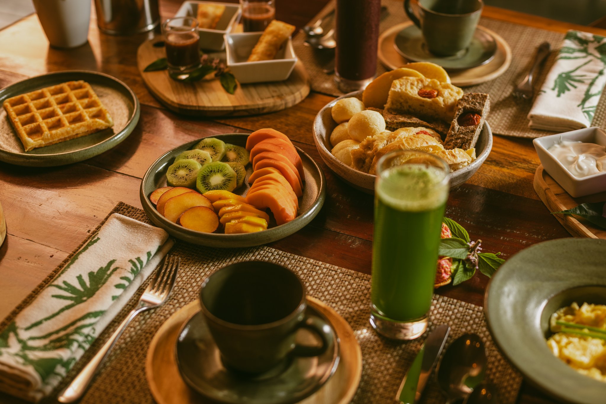 Farm-fresh mornings at Pousada Trijunção — fuel up before the wild Cerrado begins. A generous Pousada Trijunção breakfast table laid with sliced kiwi, peach and papaya, golden waffles, warm bread rolls, a vibrant green juice and a ceramic coffee mug on a woven placemat