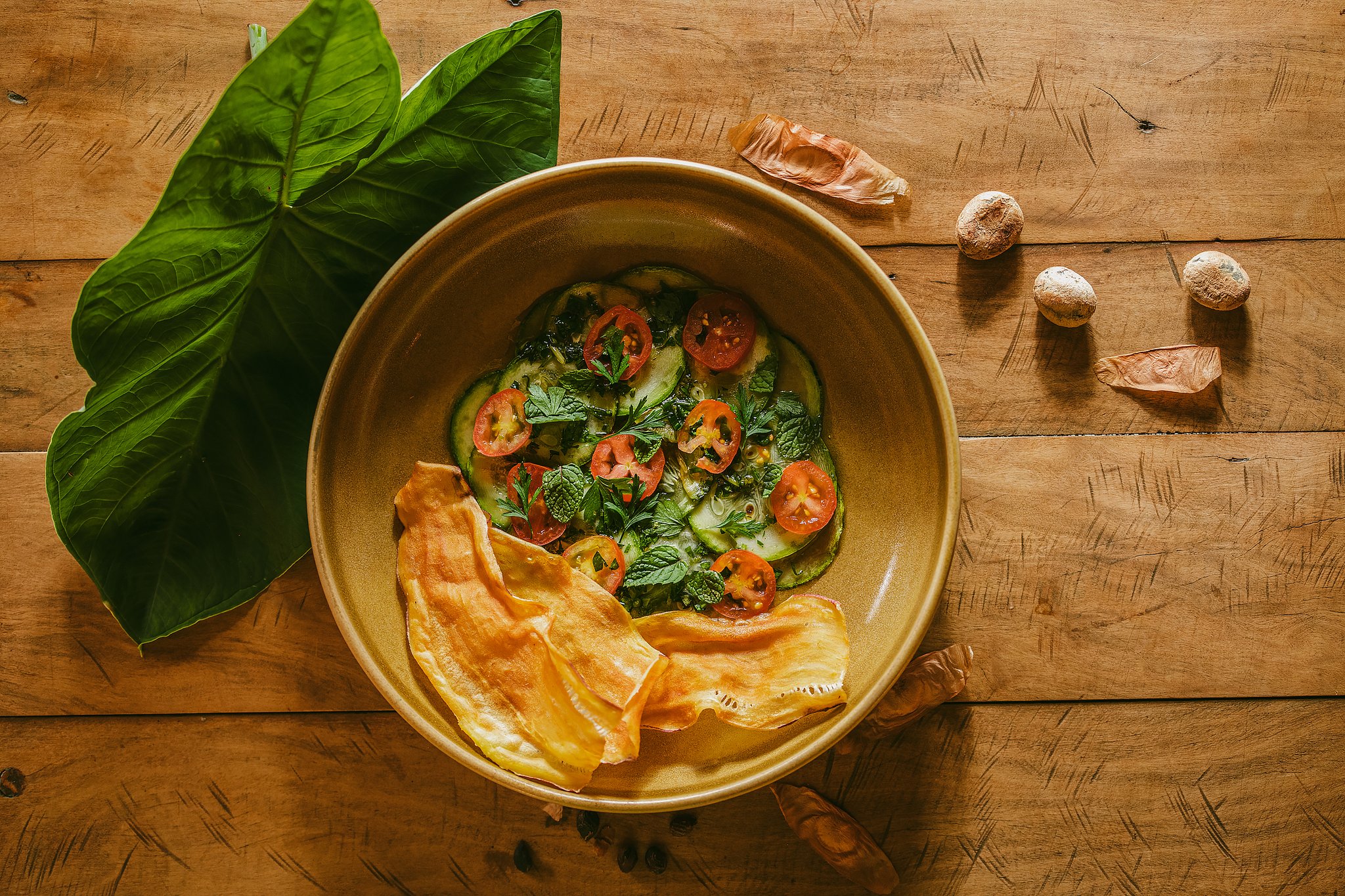 Farm-fresh flavours straight from the Cerrado's heart. Overhead view of a ceramic bowl filled with sliced zucchini, cherry tomatoes and fresh herbs served with crispy chips at Pousada Trijunção, surrounded by tropical leaves and wild seeds on a rustic wooden table