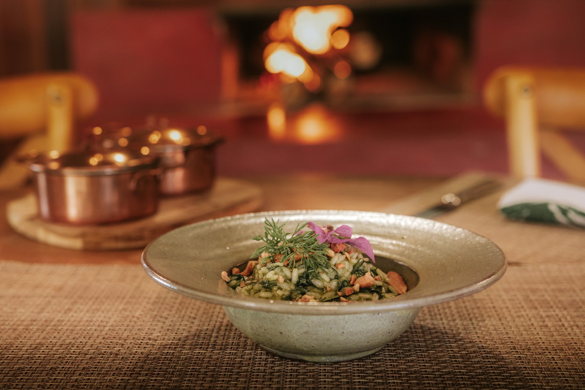 Farm-fresh flavours by the fire — dinner at Pousada Trijunção. Photo: Albori Ribeiro A handcrafted ceramic bowl of herb risotto garnished with fresh dill and a pink edible flower, served fireside at Pousada Trijunção with copper pots glowing in the warm background