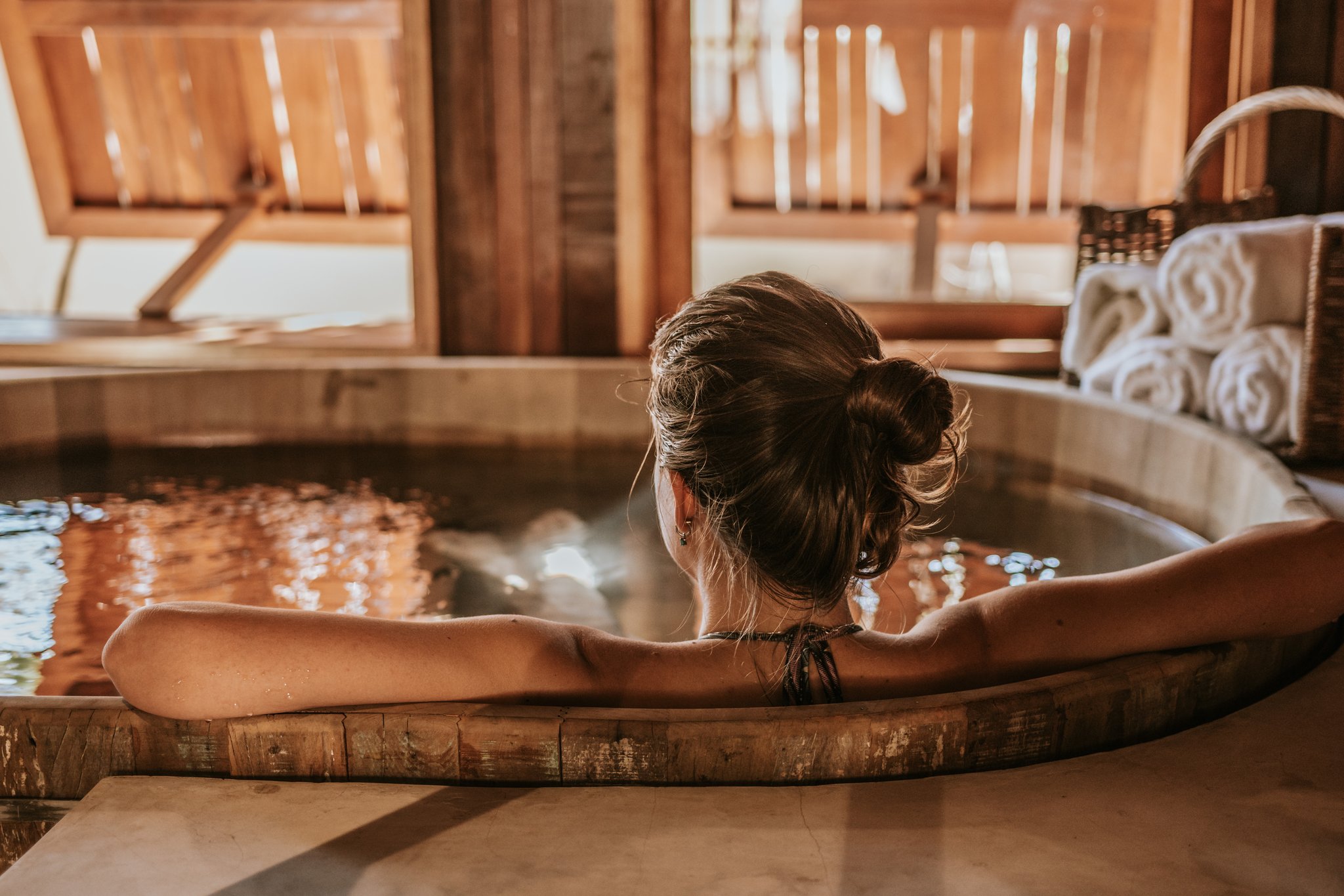 A woman soaking in a circular wooden hot tub inside a warm timber-clad room at Pousada Trijunção, with rolled white towels nearby