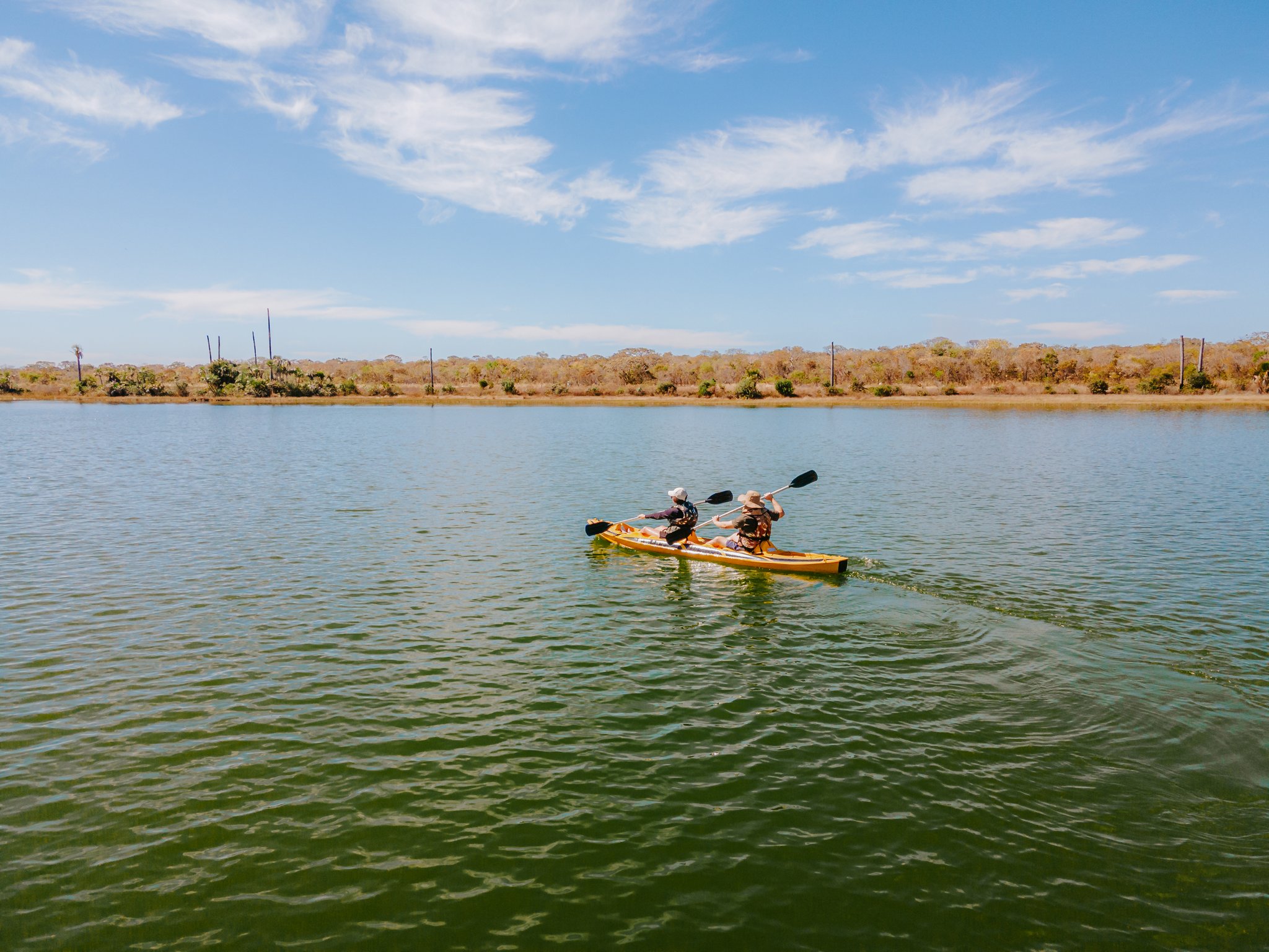 Morning on the water — paddles dipping in perfect sync at Lagoa das Araras. Two paddlers in a yellow tandem kayak gliding across the calm green waters of Lagoa das Araras at Pousada Trijunção, with dry Cerrado woodland stretching along the far shore beneath a sunny blue sky