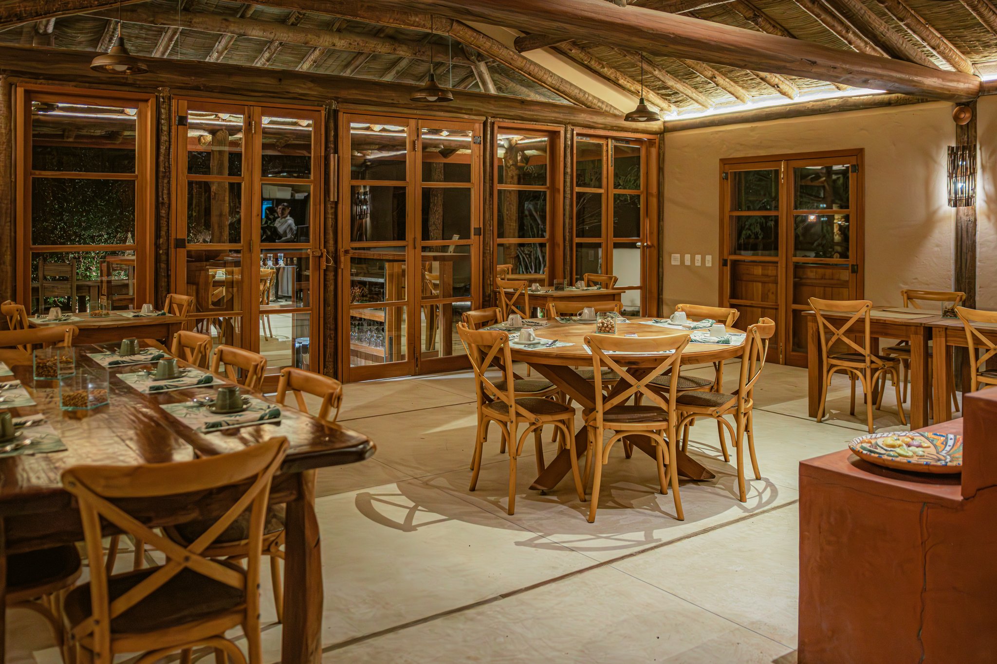 Rustic elegance meets the wild — dinner awaits at Pousada Trijunção. Photo: Albori Ribeiro Warmly lit dining room at Pousada Trijunção featuring rustic wooden cross-back chairs, round and long communal tables set with green napkins, thatched ceiling with exposed timber beams, and large wood-framed glass doors opening to the garden