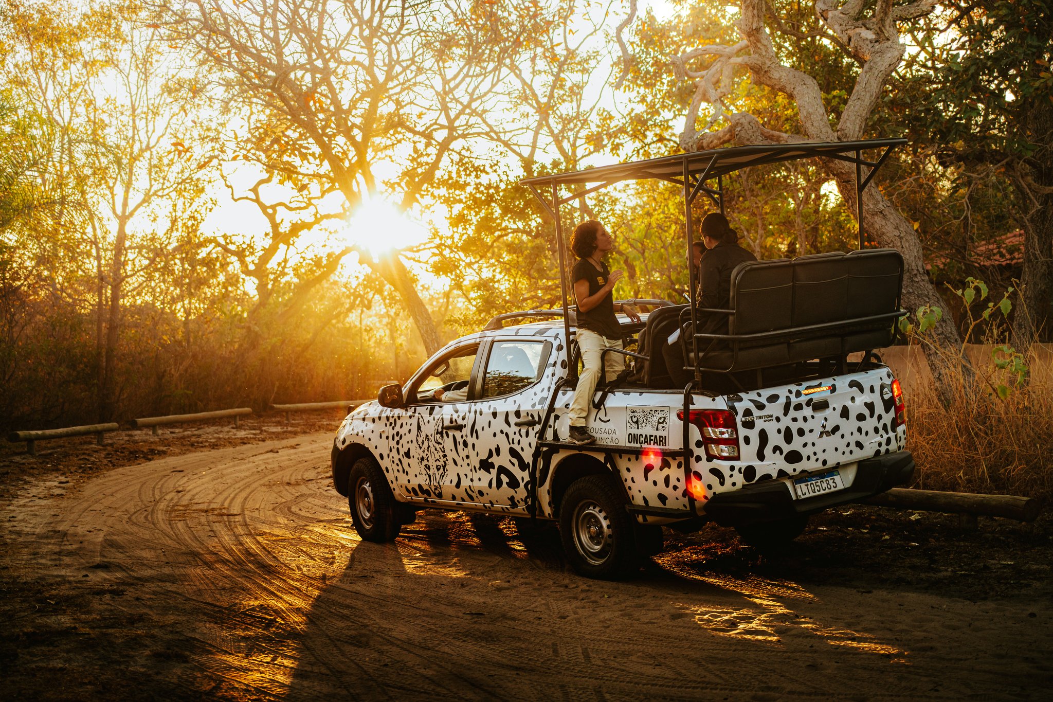 Golden hour on the trail — the iconic Onçafari vehicle soaks up the Cerrado light. Photo: Albori Ribeiro Oncafari jaguar-spotted safari truck with guests on board paused on a dirt track at golden-hour sunset in the Cerrado at Pousada Trijunção
