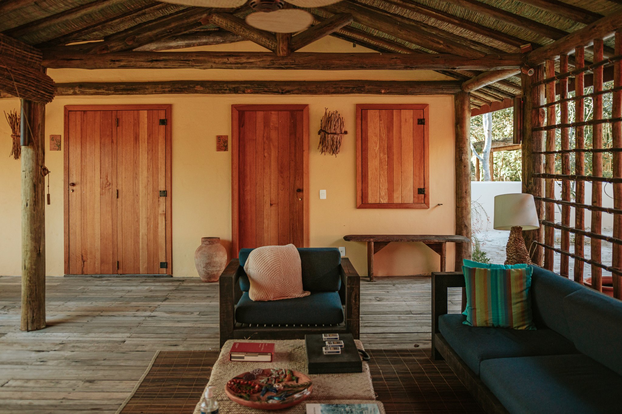 Kick back on the veranda — Cerrado views just beyond the treeline. Photo: Albori Ribeiro Open-air covered veranda at Pousada Trijunção with teal armchairs, wooden log pillars, warm terracotta walls and handcrafted wooden doors