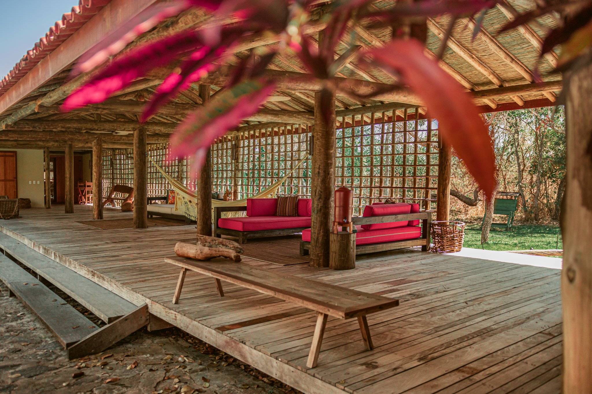Kick back on the deck — where handcrafted wood, crimson cushions and Cerrado air set the mood. Photo: Albori Ribeiro Open-air lounge at Pousada Trijunção featuring a rustic thatched roof supported by log columns, red-cushioned wooden sofas, hanging hammocks, and a wide timber deck framed by pink bougainvillea
