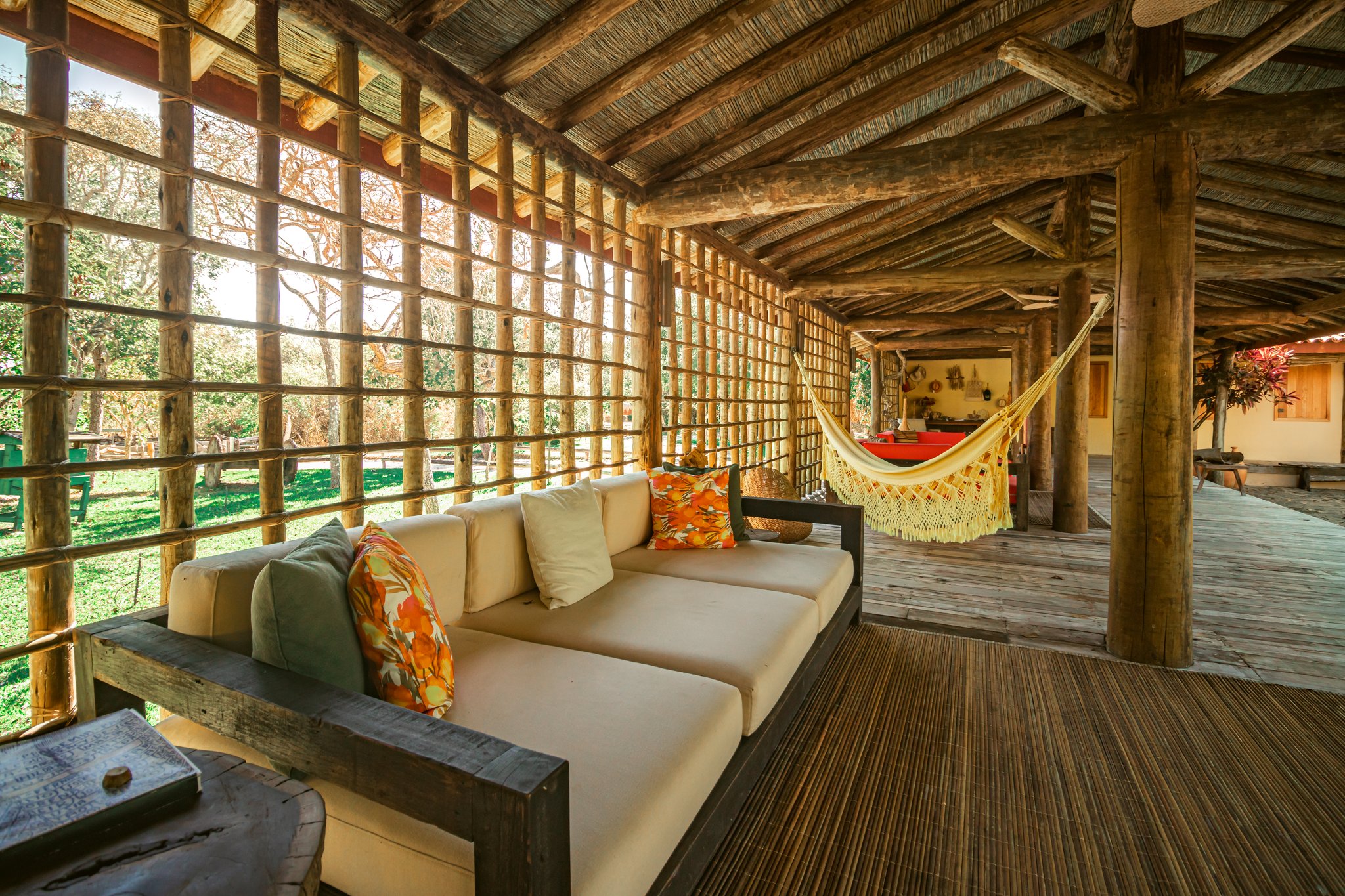 Slow down, sink in — the verandas at Pousada Trijunção were made for doing absolutely nothing. Photo: Albori Ribeiro Open-air veranda at Pousada Trijunção with a cream sofa, colorful cushions, a fringed hammock, and a rustic log-and-thatch structure overlooking lush Cerrado gardens