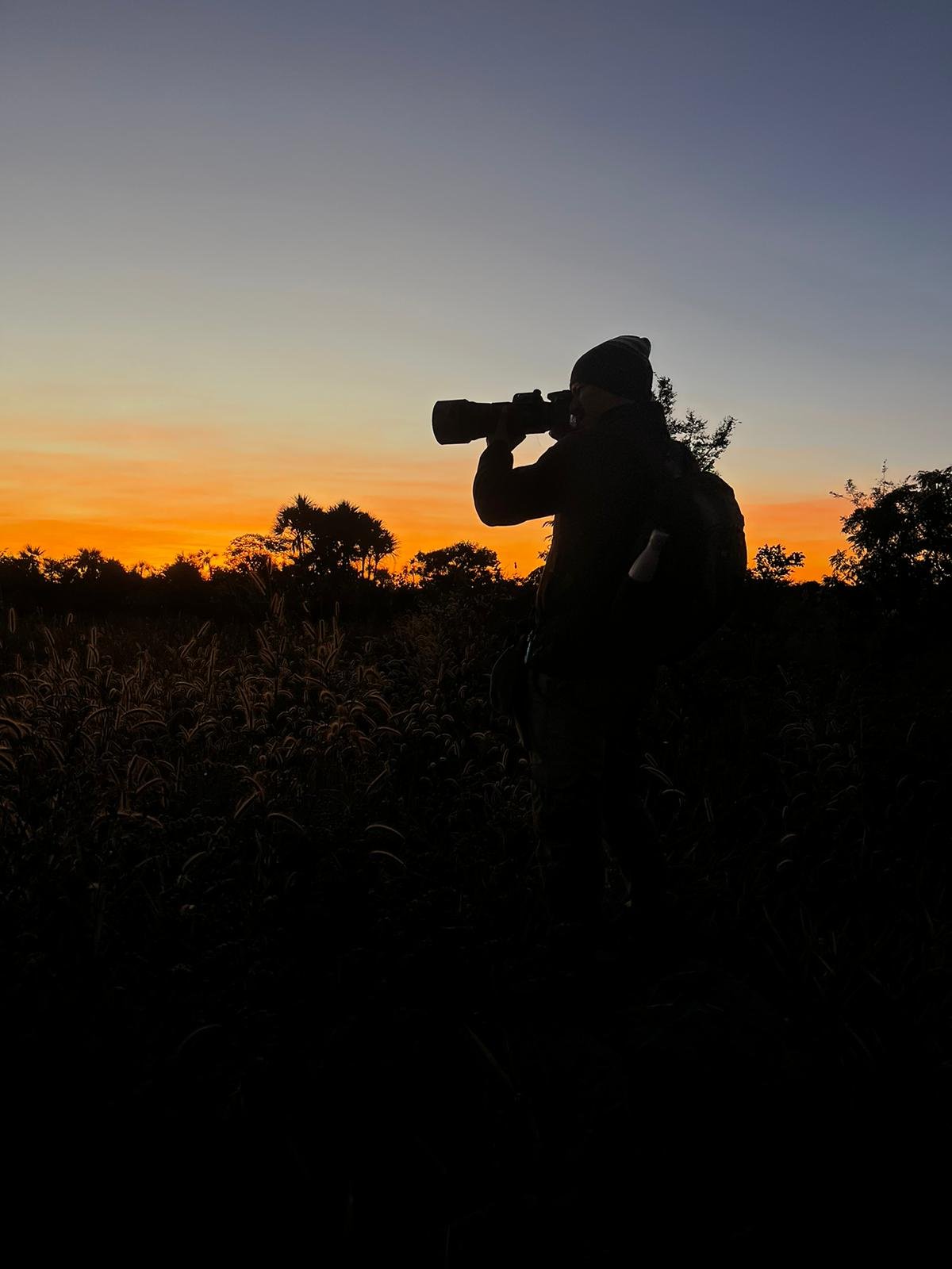 Golden hour in the Cerrado — every dawn holds a new story. Silhouette of a wildlife photographer with a telephoto lens raised to their eye, standing among tall Cerrado grasses against a vivid orange and blue sunrise sky at Pousada Trijunção
