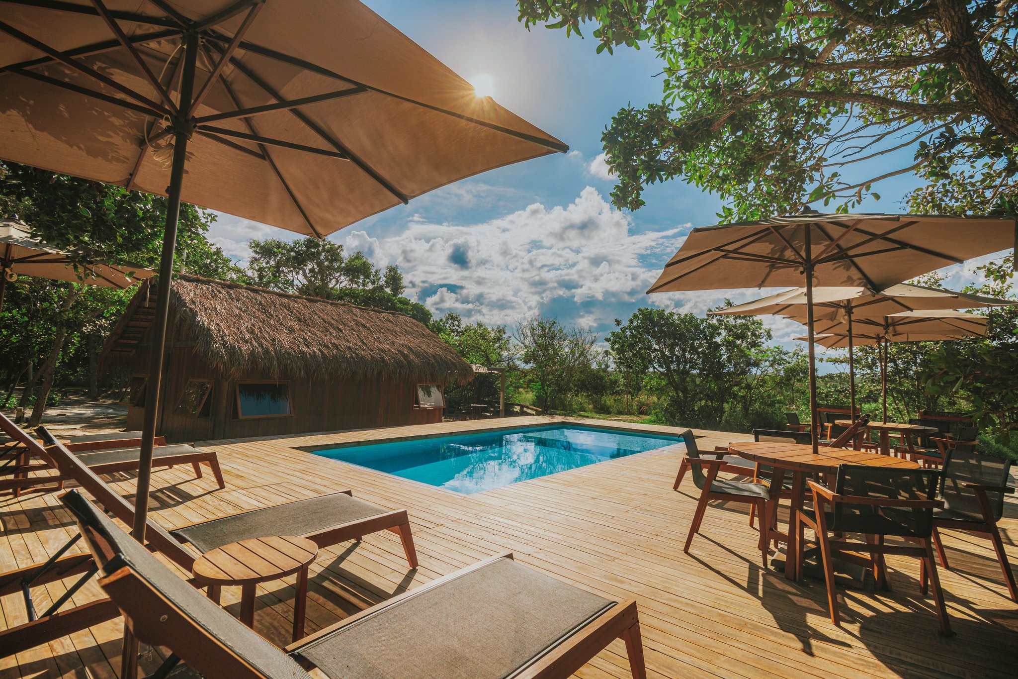 Cool off between adventures — the pool deck at Pousada Trijunção invites pure relaxation. Photo: Albori Ribeiro Sun-drenched wooden pool deck at Pousada Trijunção with lounge chairs, canvas umbrellas, a thatched-roof building, and green Cerrado vegetation under a blue sky