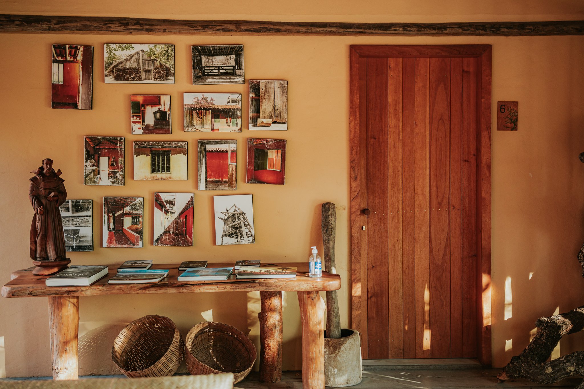 Every photo on that wall has a story the staff will happily tell you. Warm ochre wall at Pousada Trijunção decorated with a grid of photographic prints depicting traditional Cerrado architecture, beside a handcrafted wooden console table holding books, a carved monk figurine and woven baskets